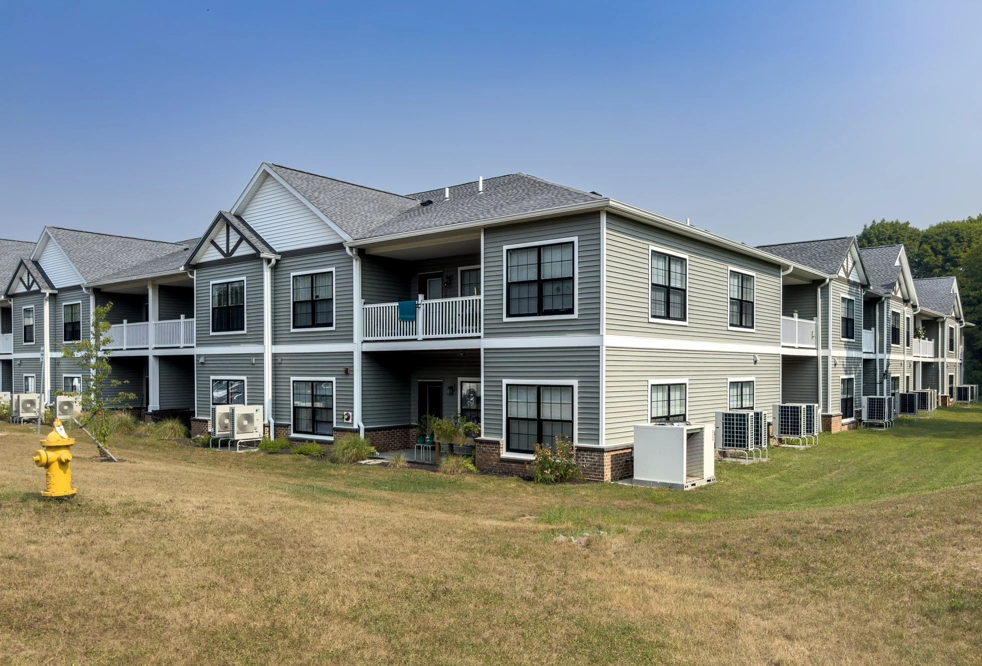 Multi-story residential building with gray siding and black window frames, small balconies, and air conditioning units outside on a well-maintained grassy area under a clear blue sky.