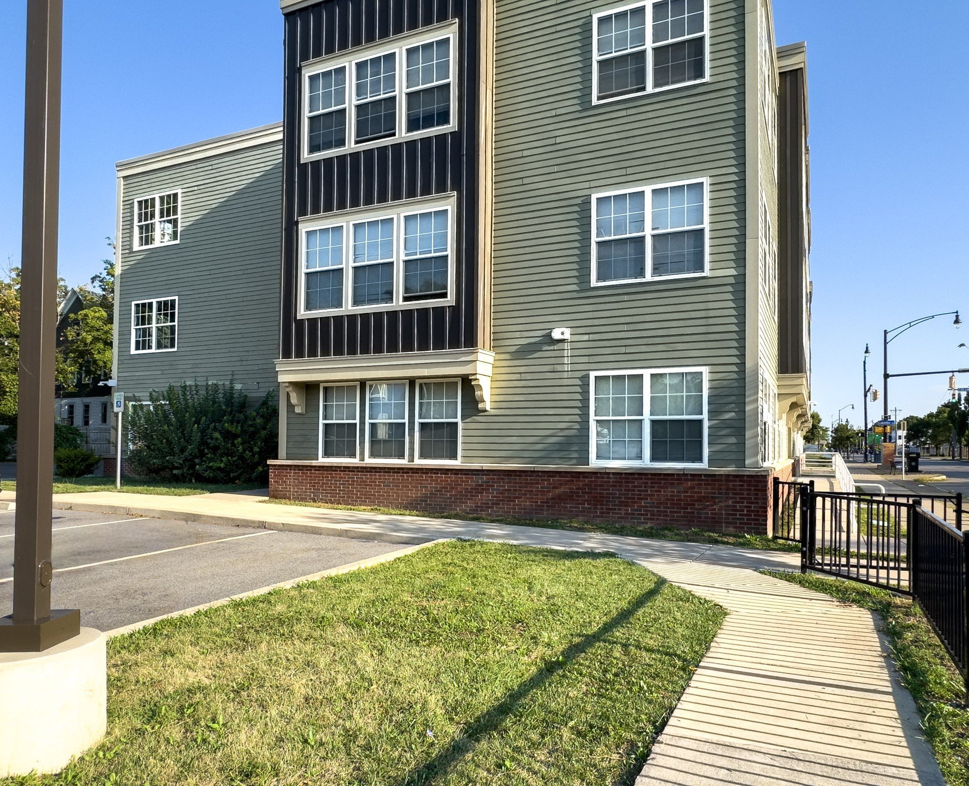 Exterior view of a multi-story apartment building with green and black siding, multiple windows, and a brick foundation. A sidewalk curves around a small grassy area with a white concrete planter and a black metal gate in the foreground. Street lamps and traffic lights are visible in the background against a clear blue sky.