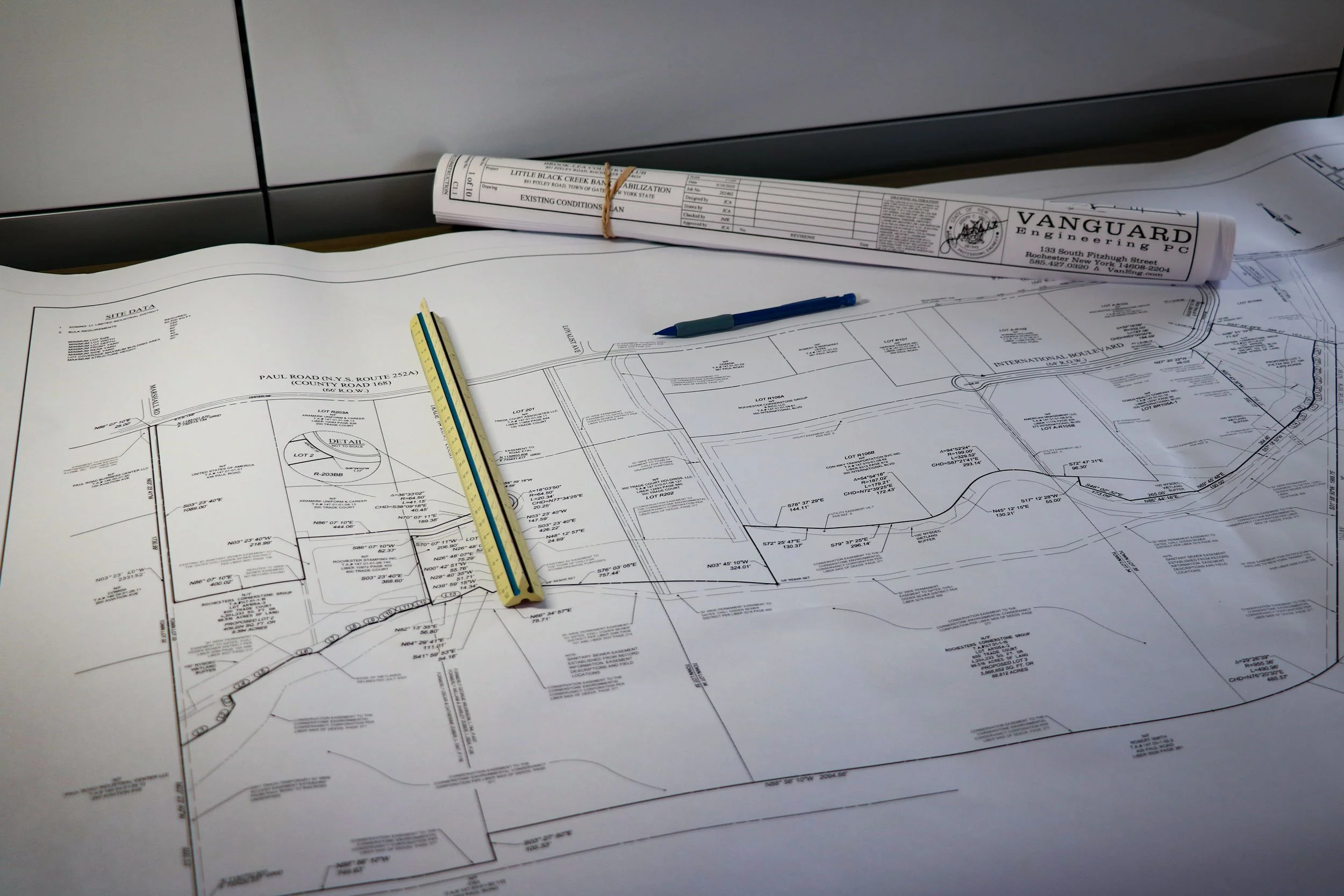 Architectural blueprint and technical drawings spread on a table with a yellow and blue ruler, a blue marker, and rolled up plans in the background.