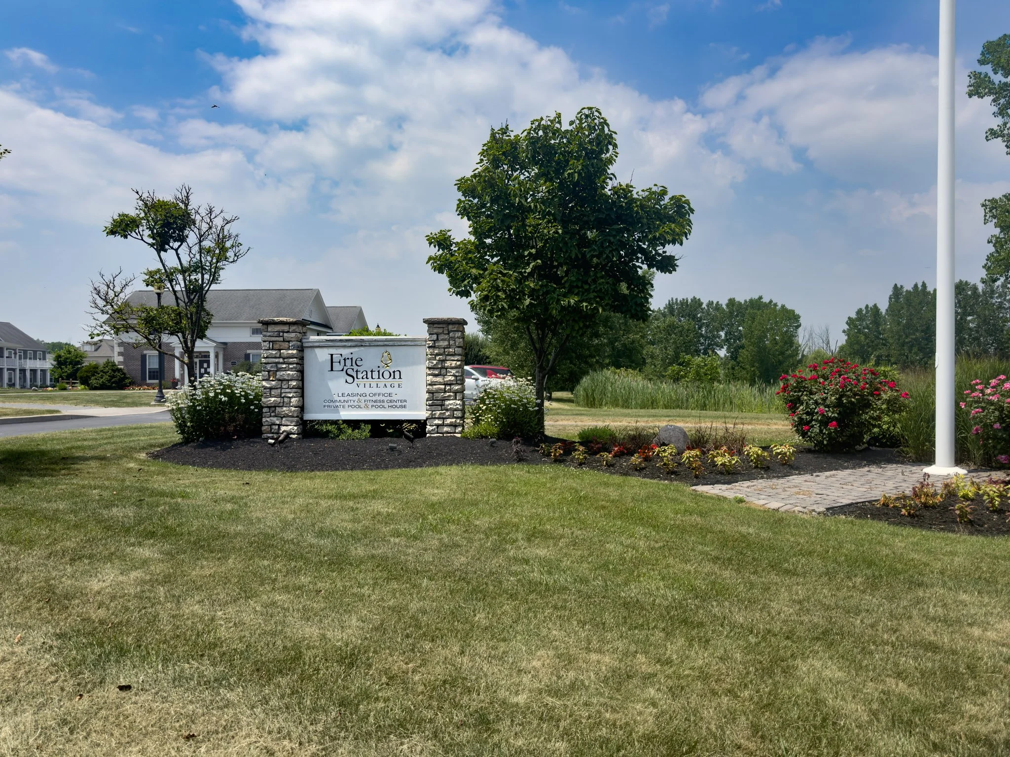 A sign for Erie Station Village community with trees, a grassy area, and houses in the background under a partly cloudy sky.