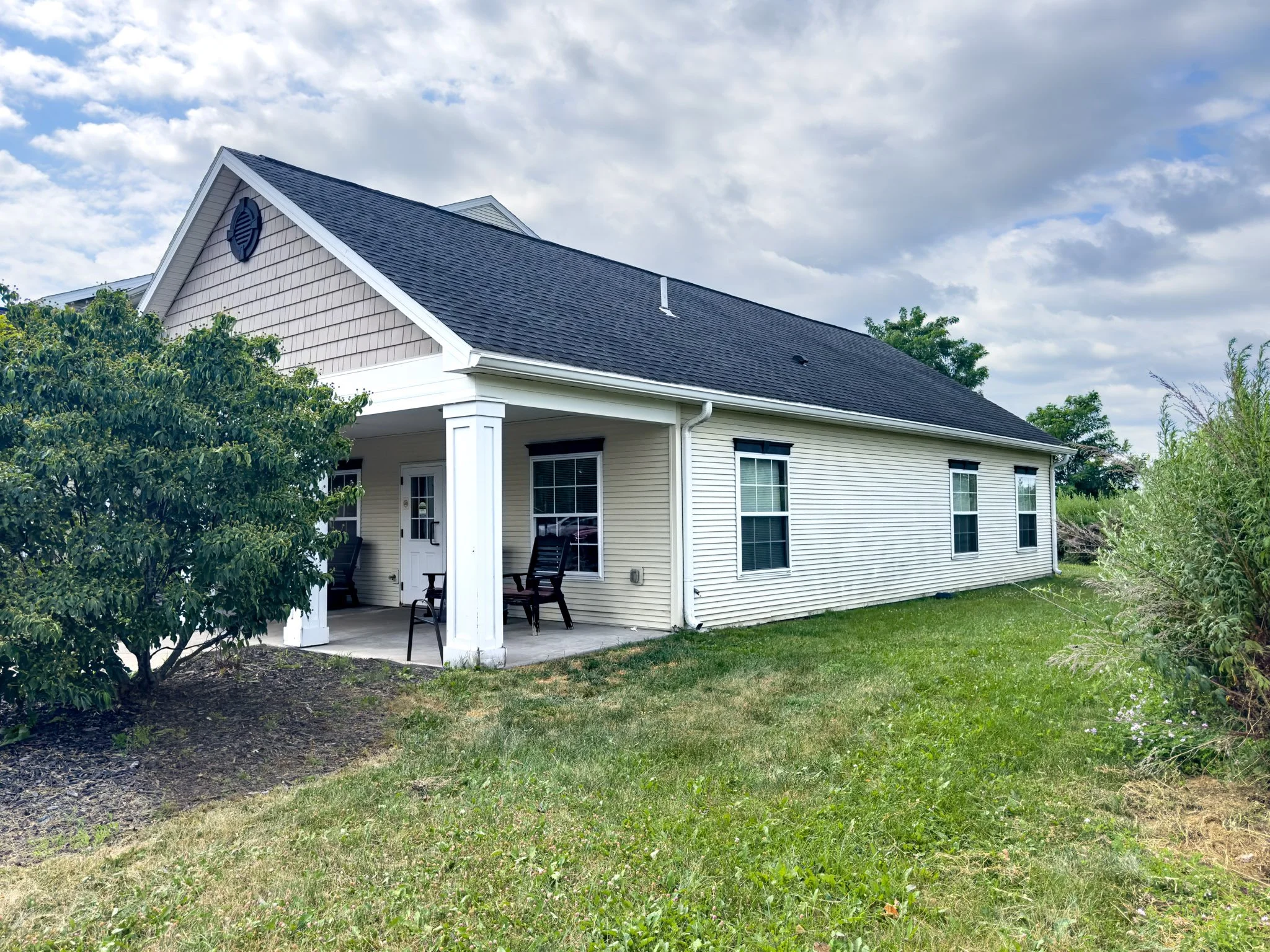 A two-story house with beige siding, white trim, and a dark shingled roof. There is a covered porch with black chairs. The yard is green with shrubs and trees, and there are clouds in the sky.
