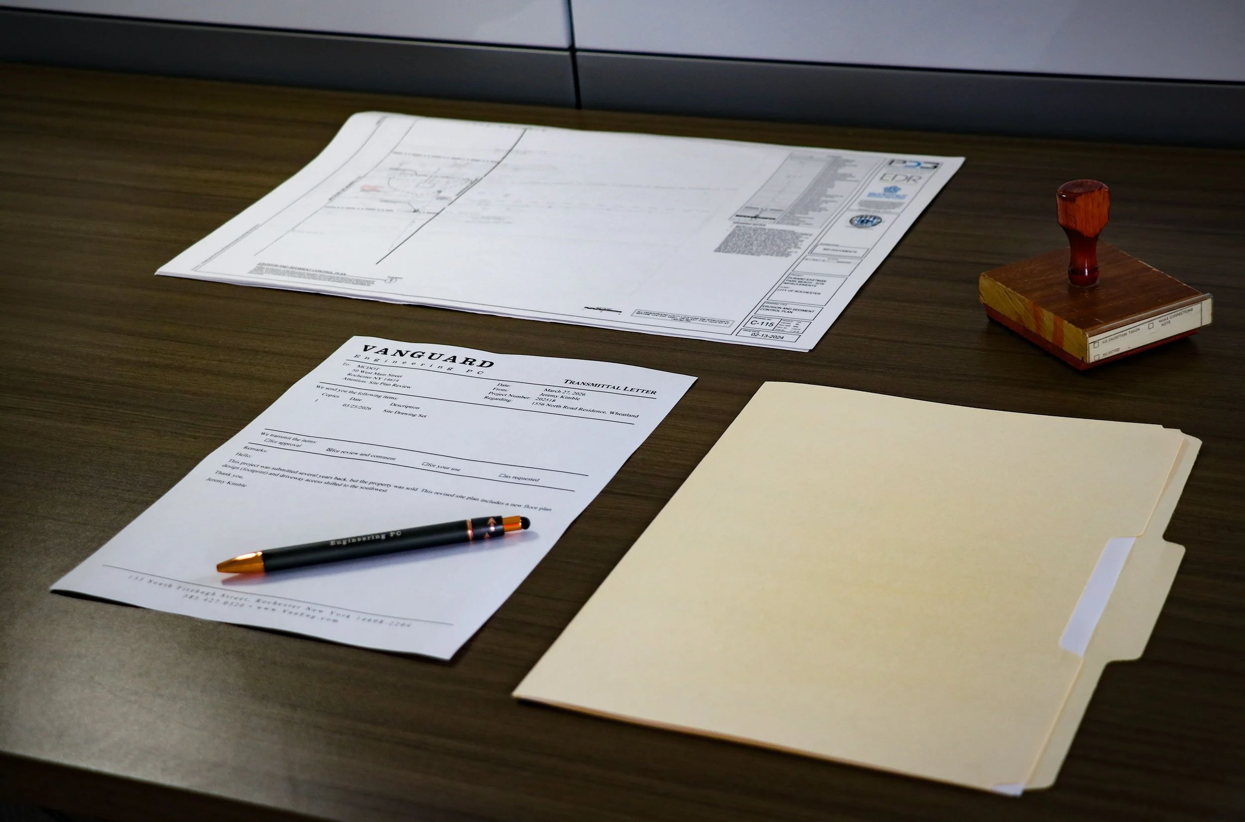 A wooden desk with architectural drawings, a business letter from Vanguard Engineering PC, a black and gold pen, and a stack of cream-colored folders.