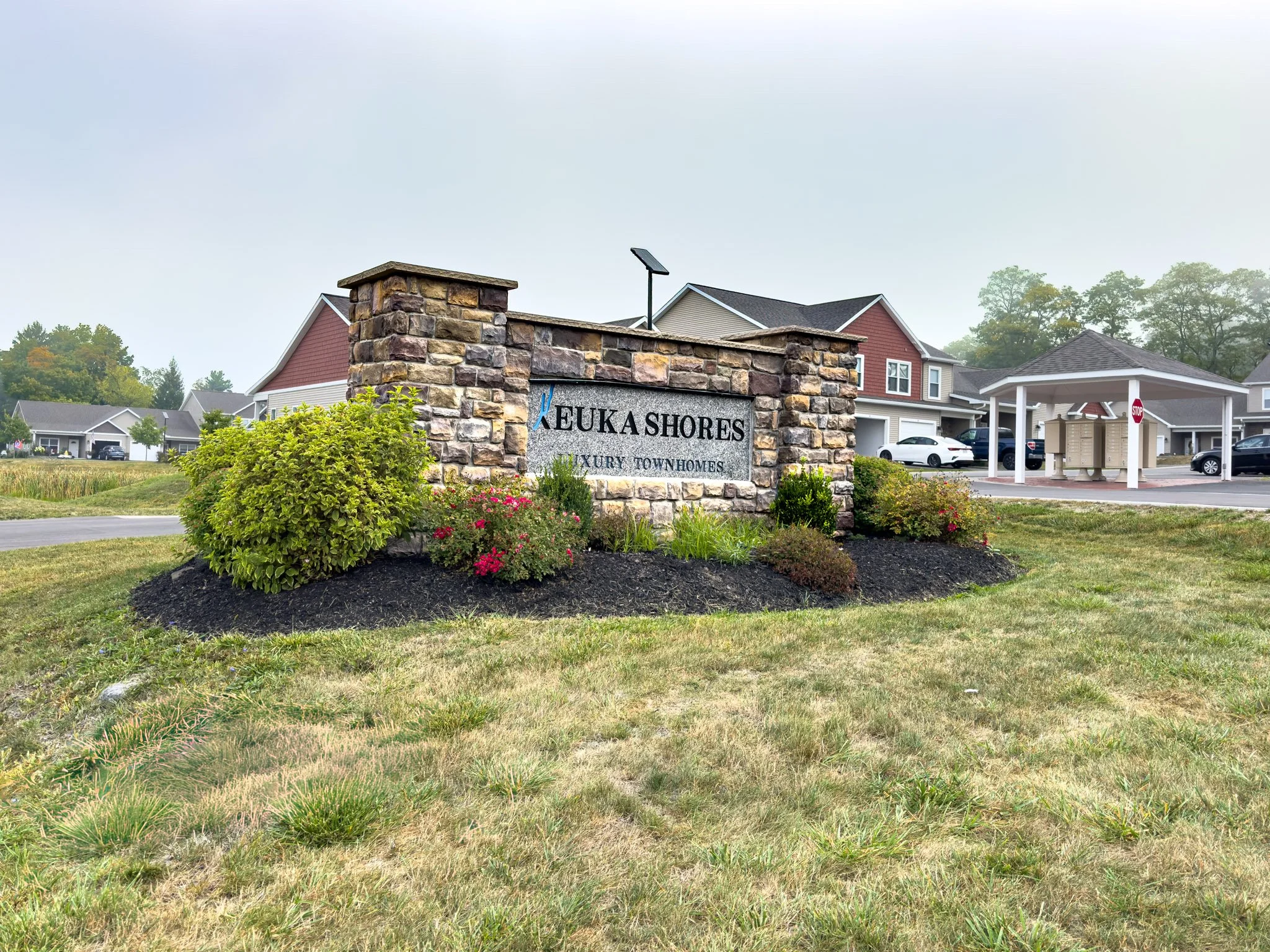 Entrance sign reading 'Keuka Shores Luxury Townhomes' at a residential community with landscaped garden, stone pillars, and modern houses in the background.