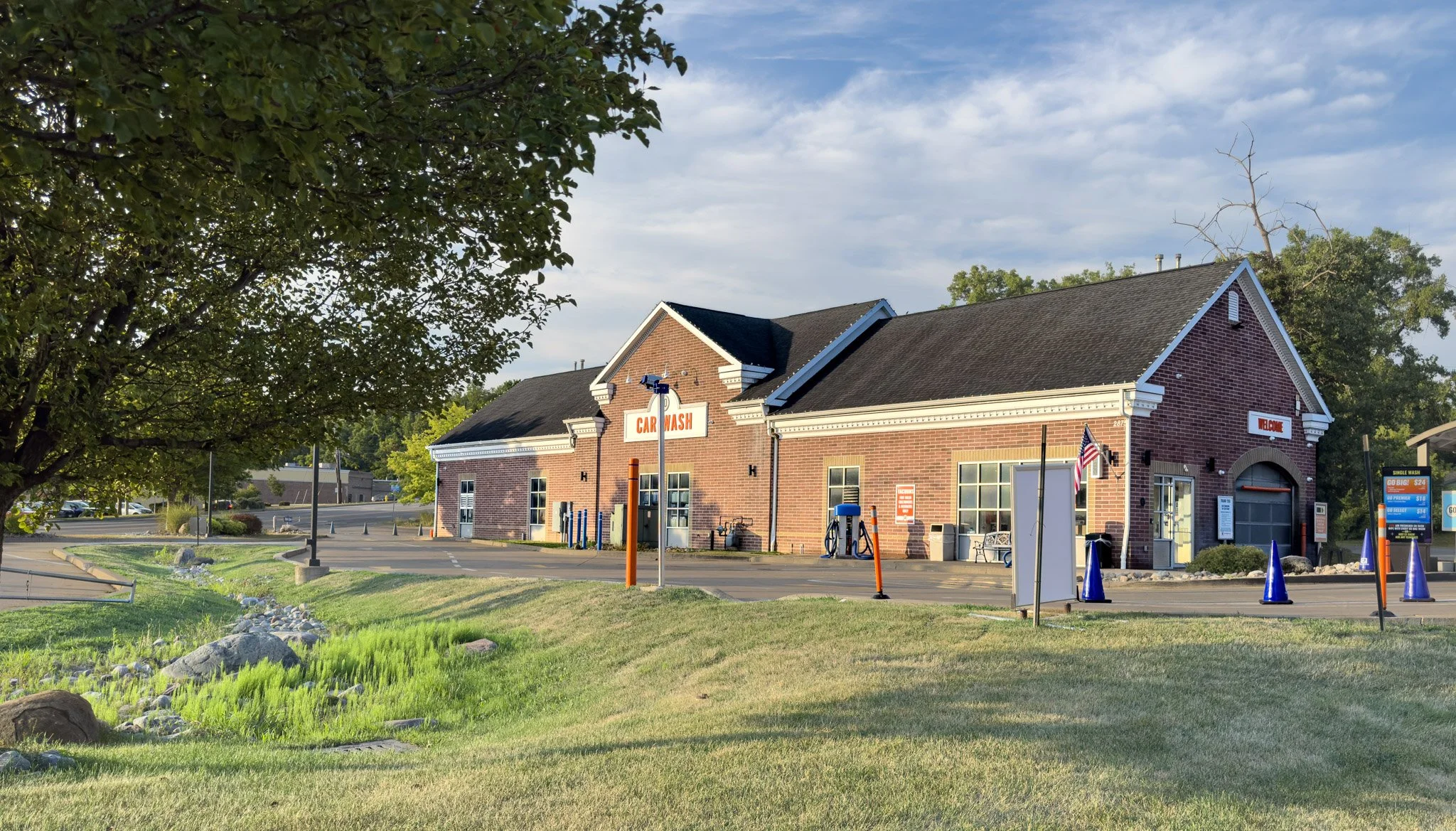 A brick building with a sign that reads 'Car Wash' and a smaller sign that says 'Welcome.' The building has a black shingle roof, windows, and outdoor washing equipment. There are parking spaces with orange cones and blue traffic cones nearby, as well as a flagpole with an American flag. The scene is outdoors on a sunny day with a partly cloudy sky and green trees in the background.