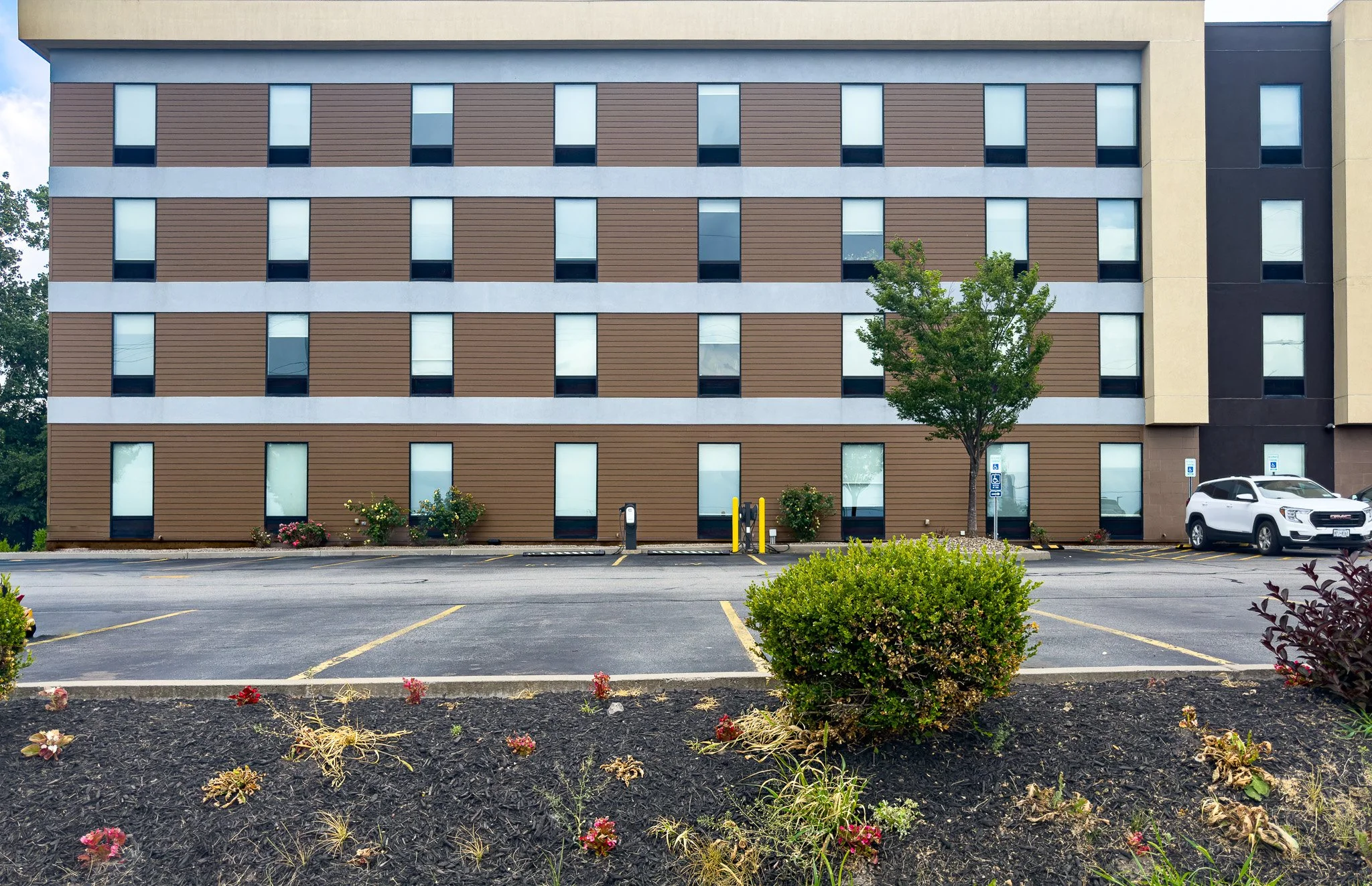 Front view of a multi-story apartment building with modern design, brown and beige exterior walls, and multiple windows. In front, there is a parking lot with a white SUV and electric vehicle charging stations. Small landscaped area with bushes, flowers, and a single tree.