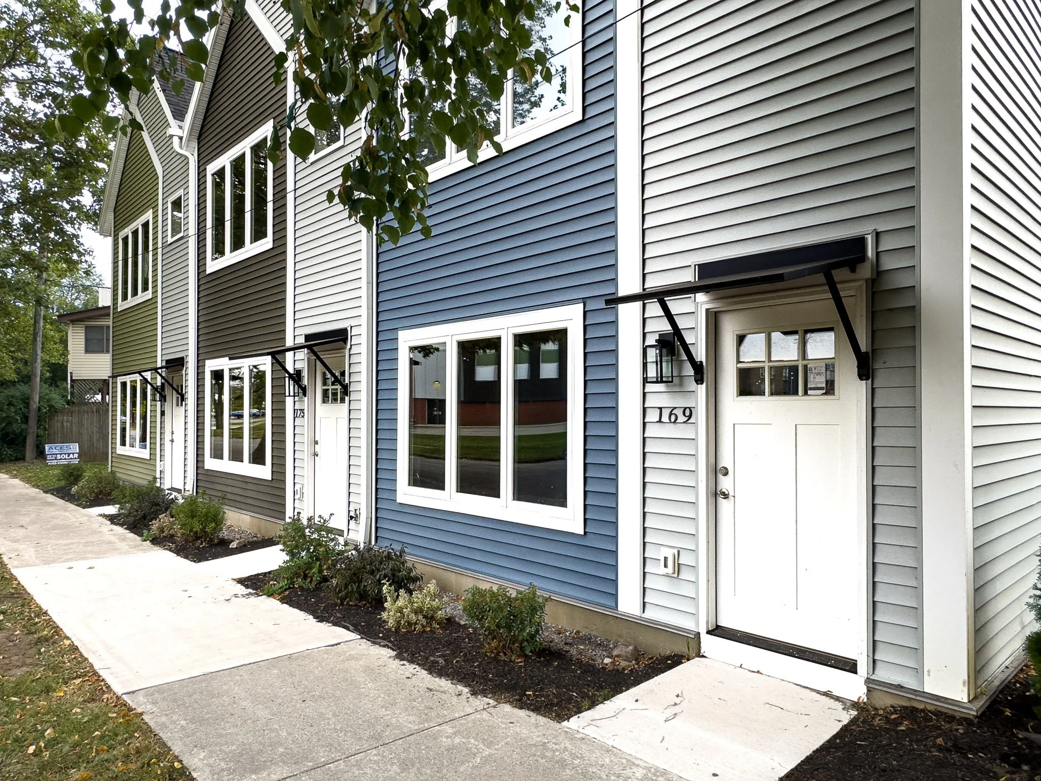 The exterior of a modern, multi-unit townhouse with blue and gray siding, white window frames, small front gardens, and a concrete sidewalk