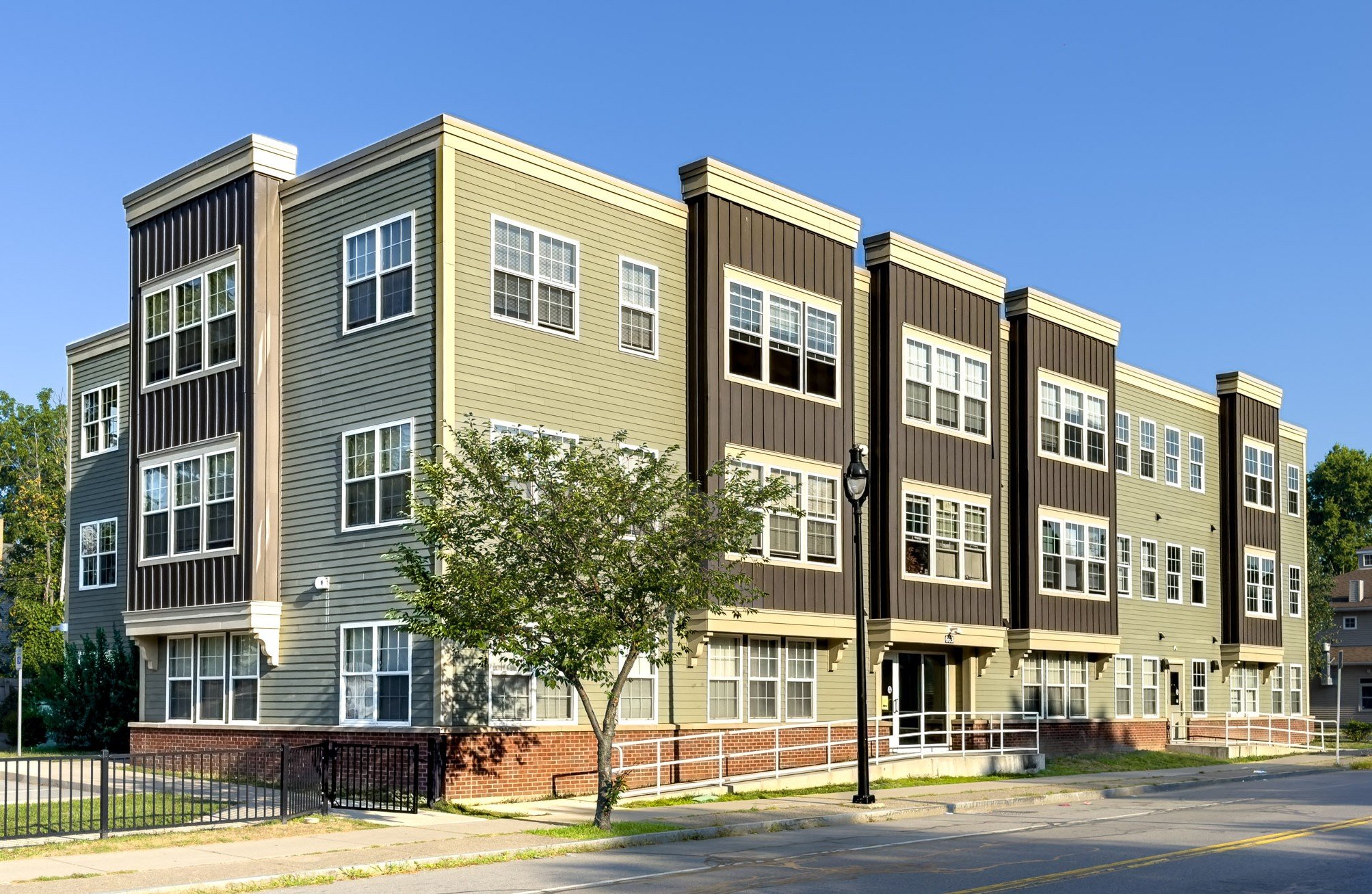 Multi-story residential building with beige and dark brown siding, multiple windows, trees, and a sidewalk in the foreground under a clear blue sky.
