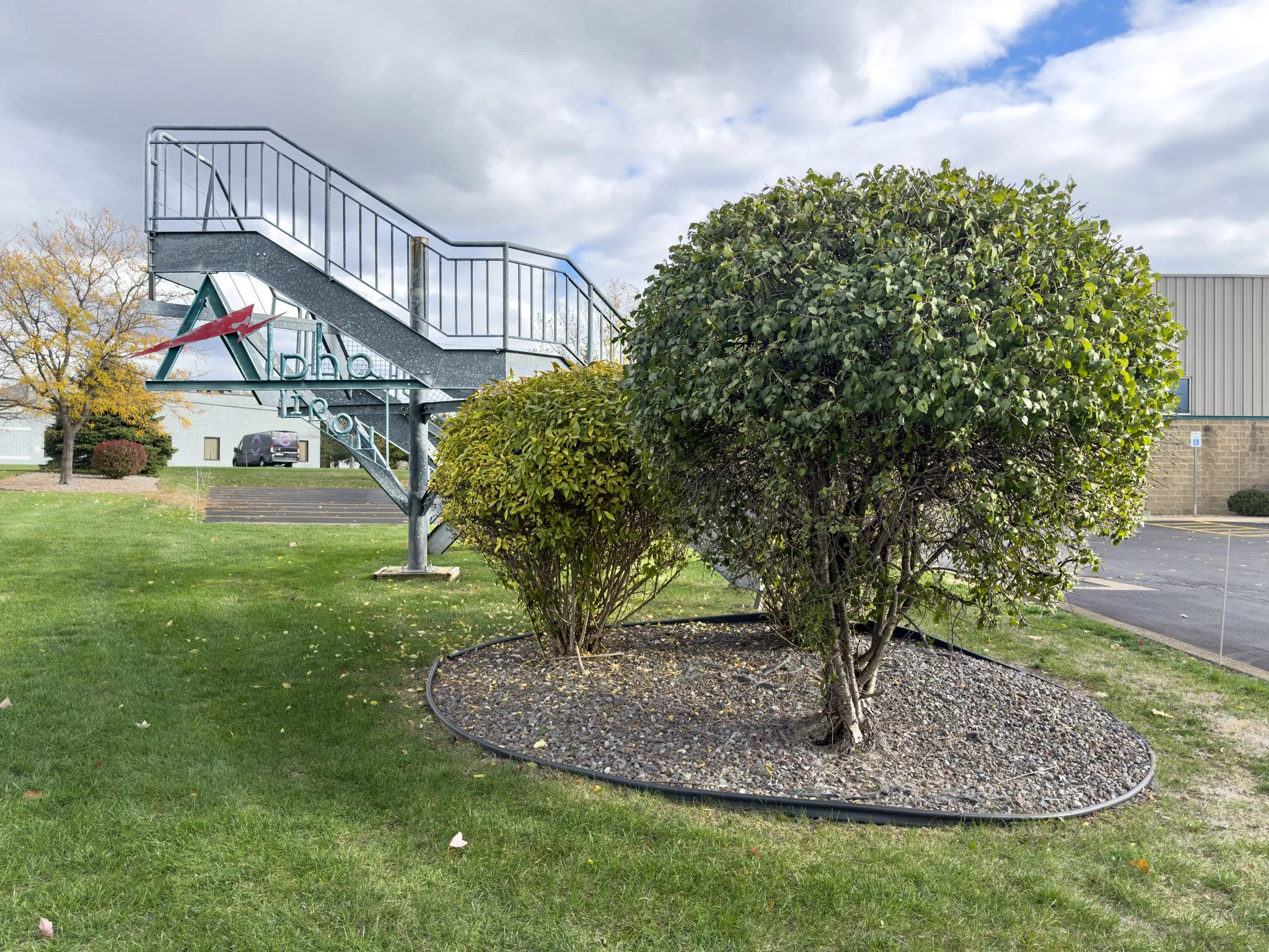 A landscaped outdoor area with a small tree surrounded by mulch, a large bush, and a grassy lawn. In the background, there is a staircase with a sign that reads 'Alpha Lion' and a parking lot with a vehicle. The sky above is cloudy with patches of blue.