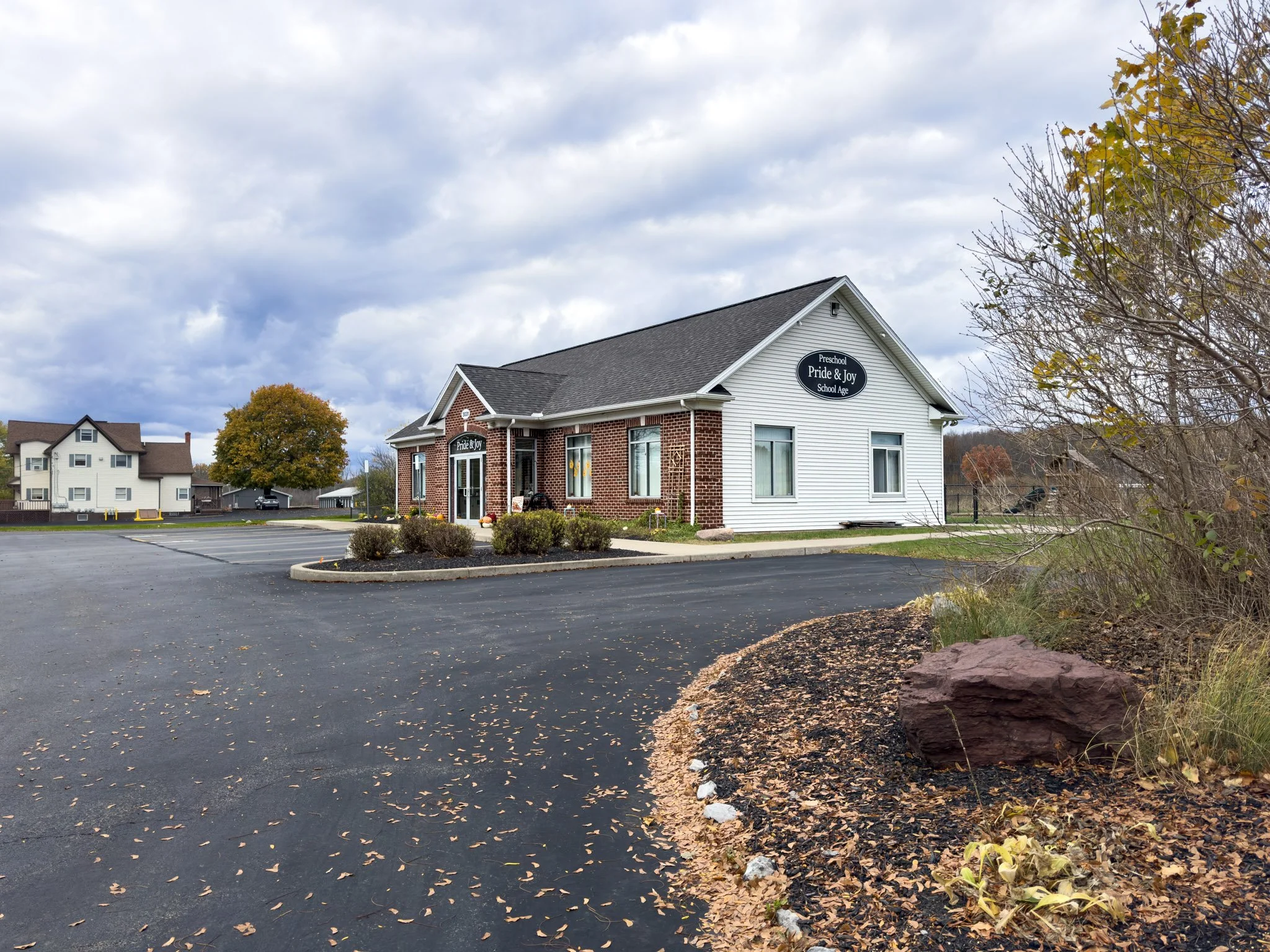 A building with a sign that reads 'Preschool Pride & Joy, School Age' located in a parking lot. The sky is cloudy, and there are trees with autumn leaves nearby.