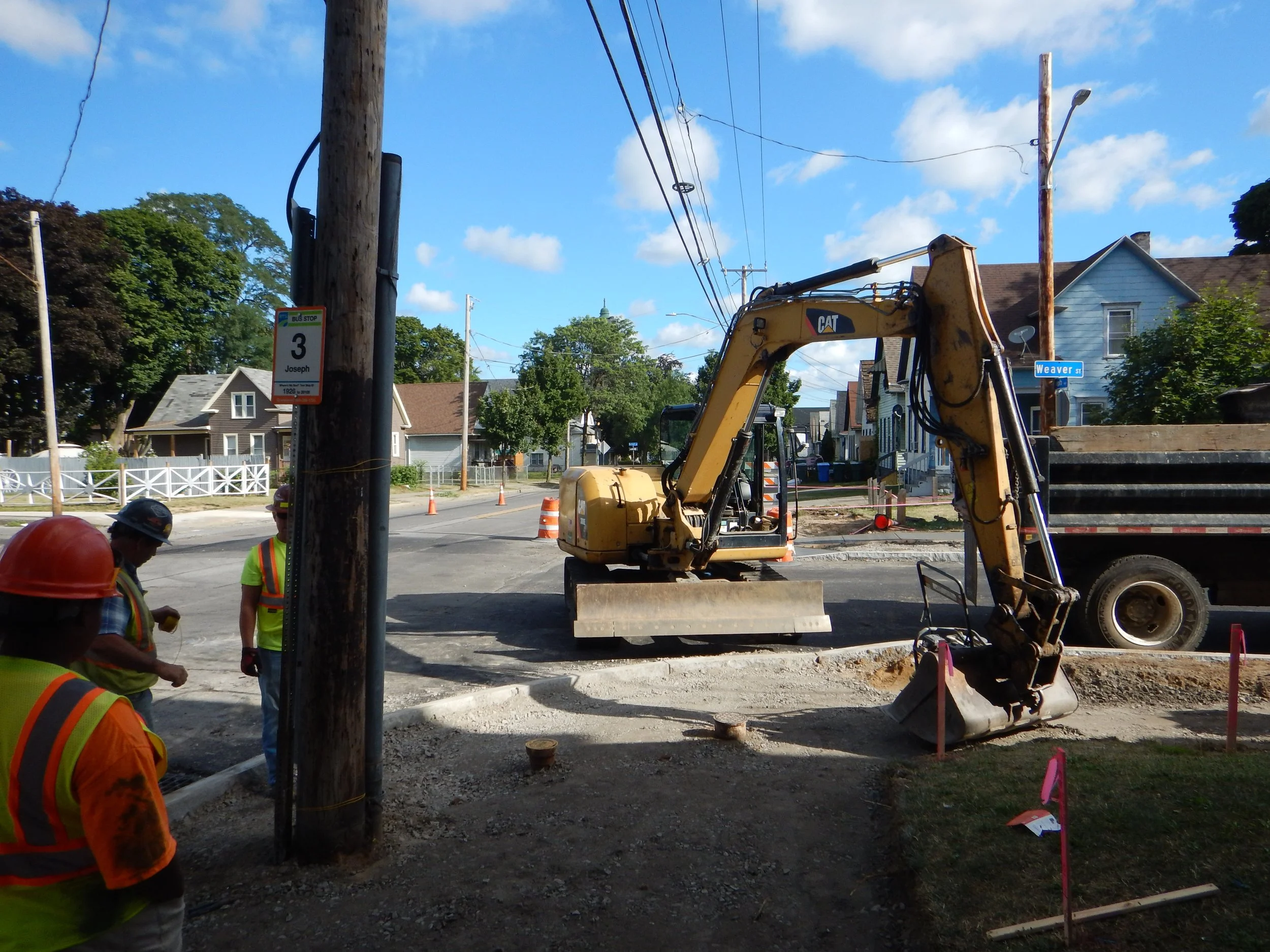 Construction workers wearing safety vests and helmets working on a street repair project. Heavy machinery and utility poles are visible, with houses and trees in the background under a blue sky.