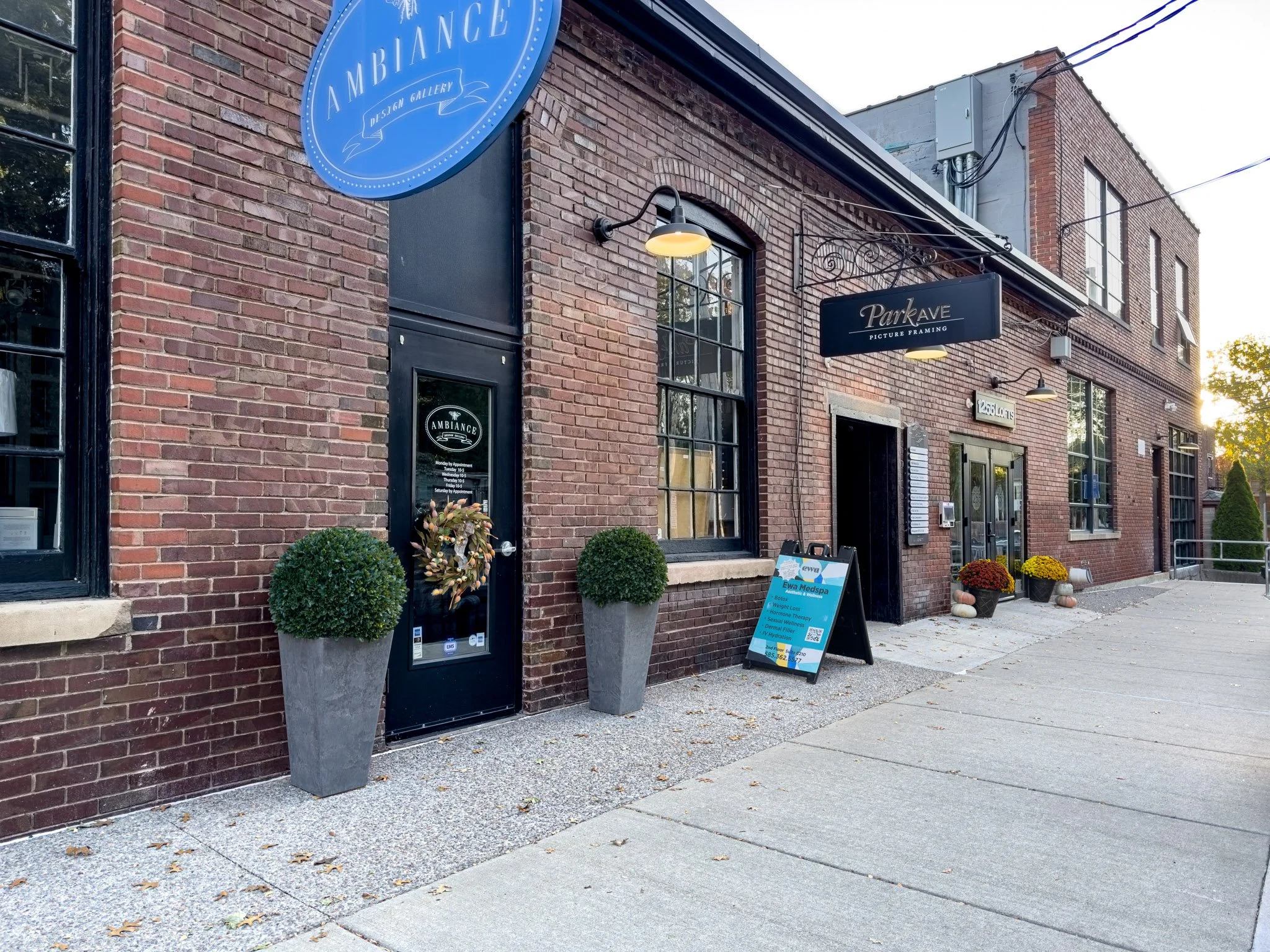 Brick storefront with large windows, displaying signs for Ambiance Design Gallery and Park Ave Picture Framing. Two large potted shrubs flank the entrance, and a sidewalk sign advertises services. There are flowers in pots outside, and the building has multiple windows on upper floors. The sky is slightly cloudy with the sun setting behind the building.