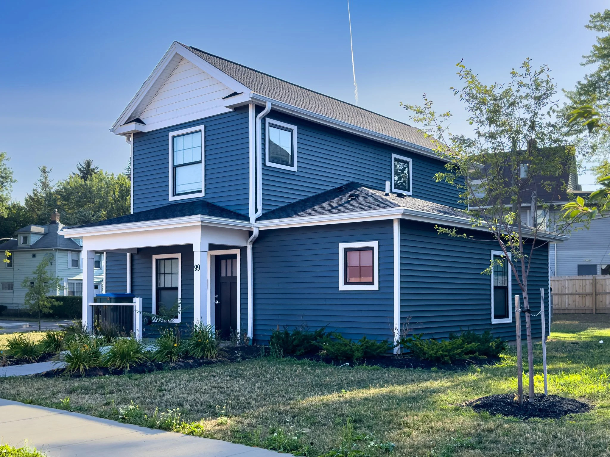 A two-story blue house with white trim, multiple windows, and a front porch, surrounded by a landscaped yard with grass, plants, and young trees, against a clear blue sky.