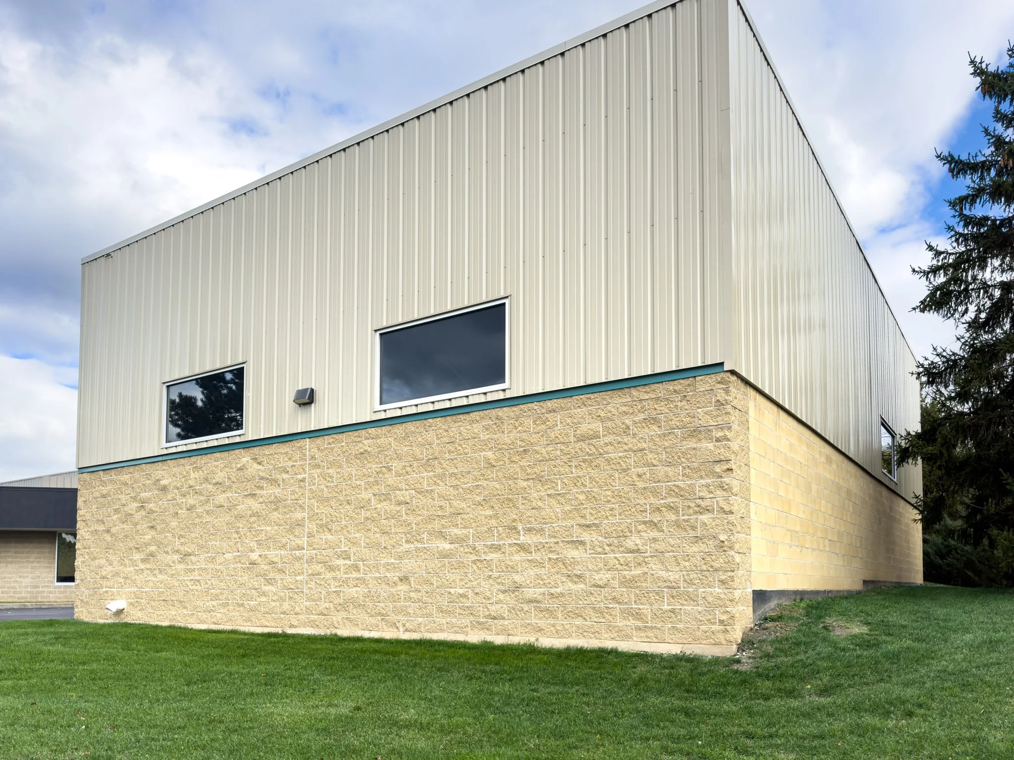 A two-story building with beige brick on the lower level and cream metal siding on the upper level, featuring two rectangular windows, set against a partly cloudy sky with a tree on the right side and a patch of green grass in the foreground.