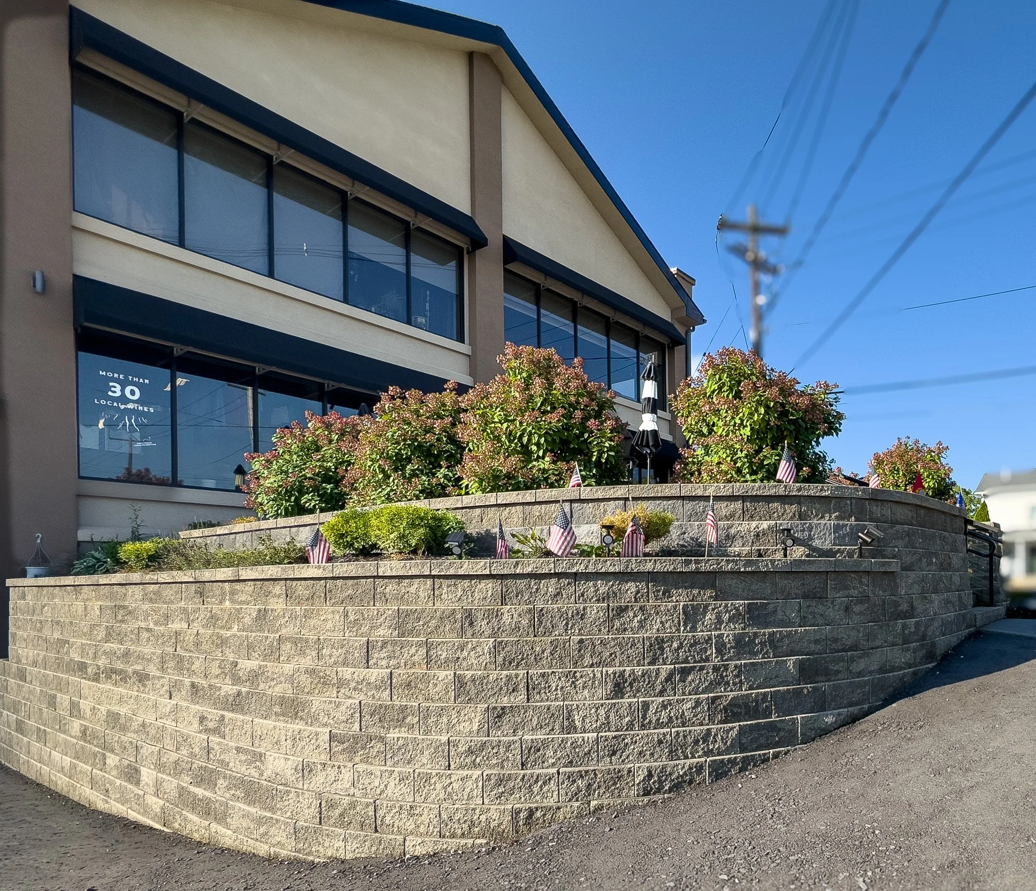 A modern multi-story building with glass windows and dark framing, sitting on a raised level with a concrete block retaining wall in front. In the foreground, there is a landscaped area with flowering bushes, small American flags, and flags of other countries. To the right, a utility pole with wires connects to the upper part of the image against a clear blue sky.
