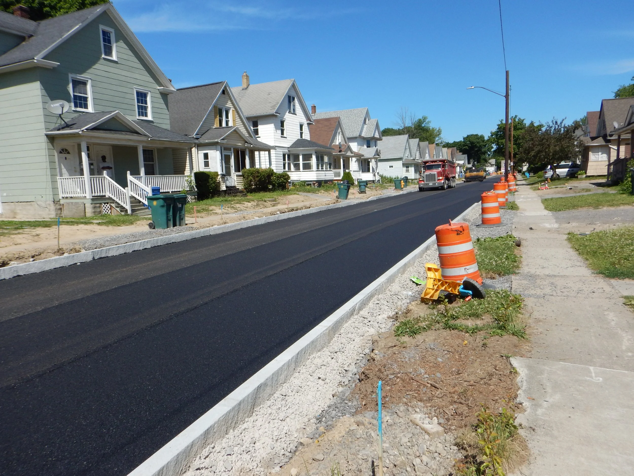 Residential street under construction with new asphalt pavement, construction cones, and houses on both sides, blue sky overhead.