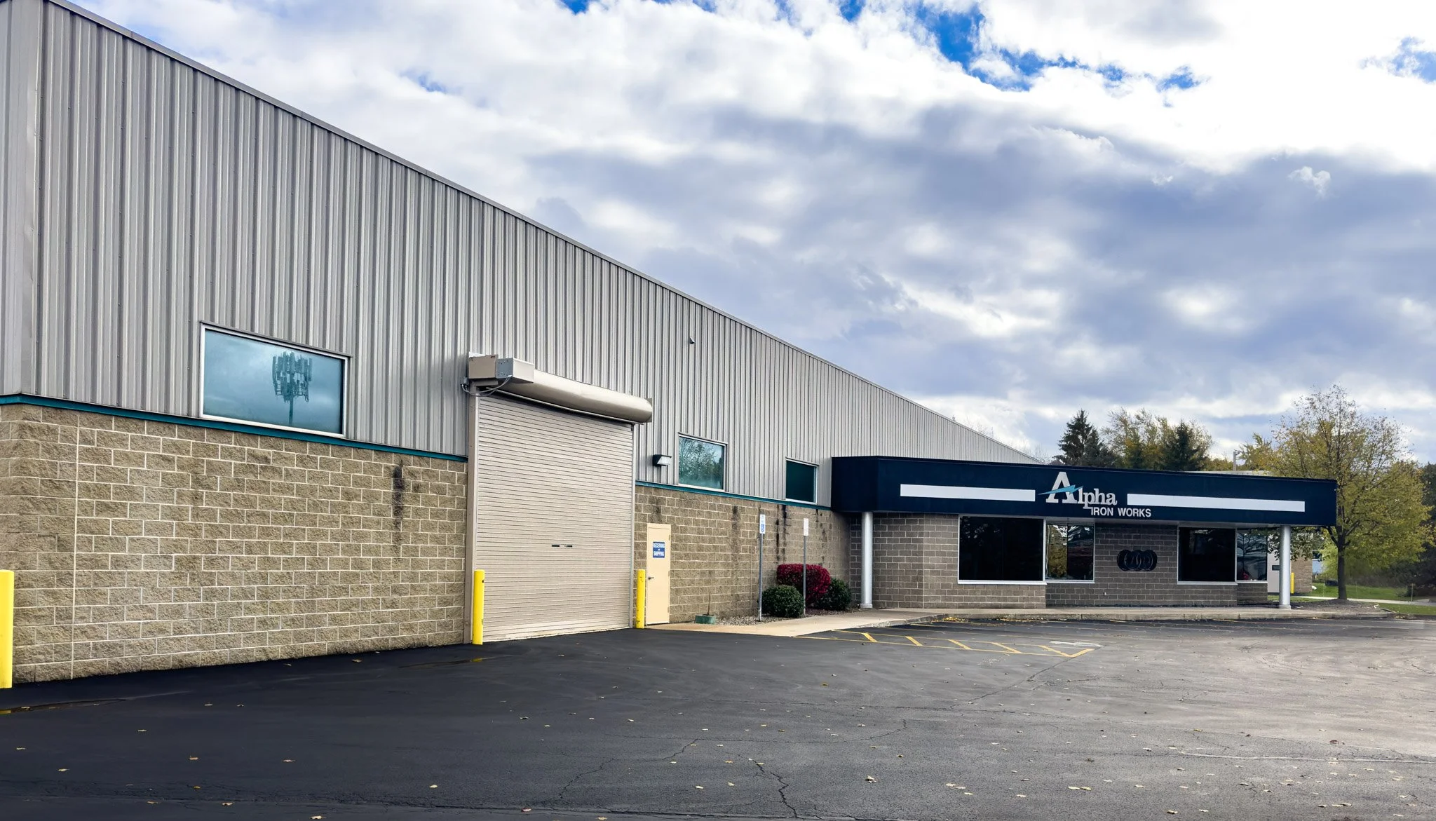 Exterior of a commercial building with a sign reading 'Alpha Iron Works', a large roller door, small window, and a parking lot in front, with trees and cloudy sky in the background.