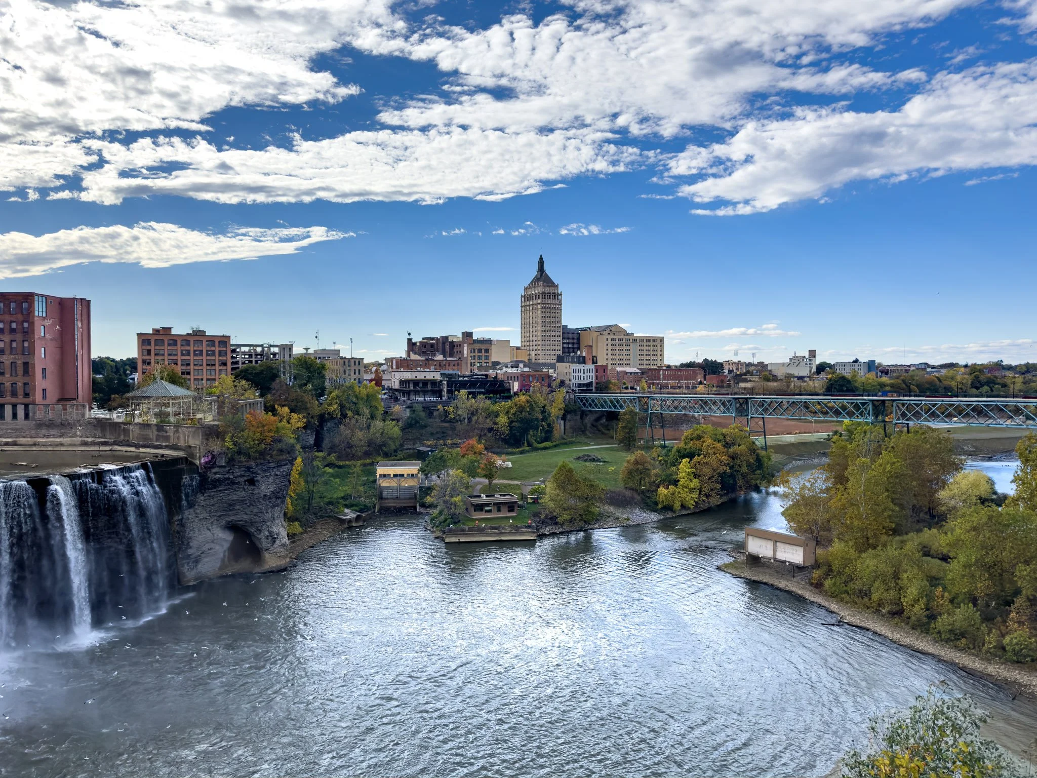 A cityscape featuring a river with a waterfall on the left, surrounded by buildings and a bridge in the foreground, with a prominent tall building in the background under a partly cloudy sky.