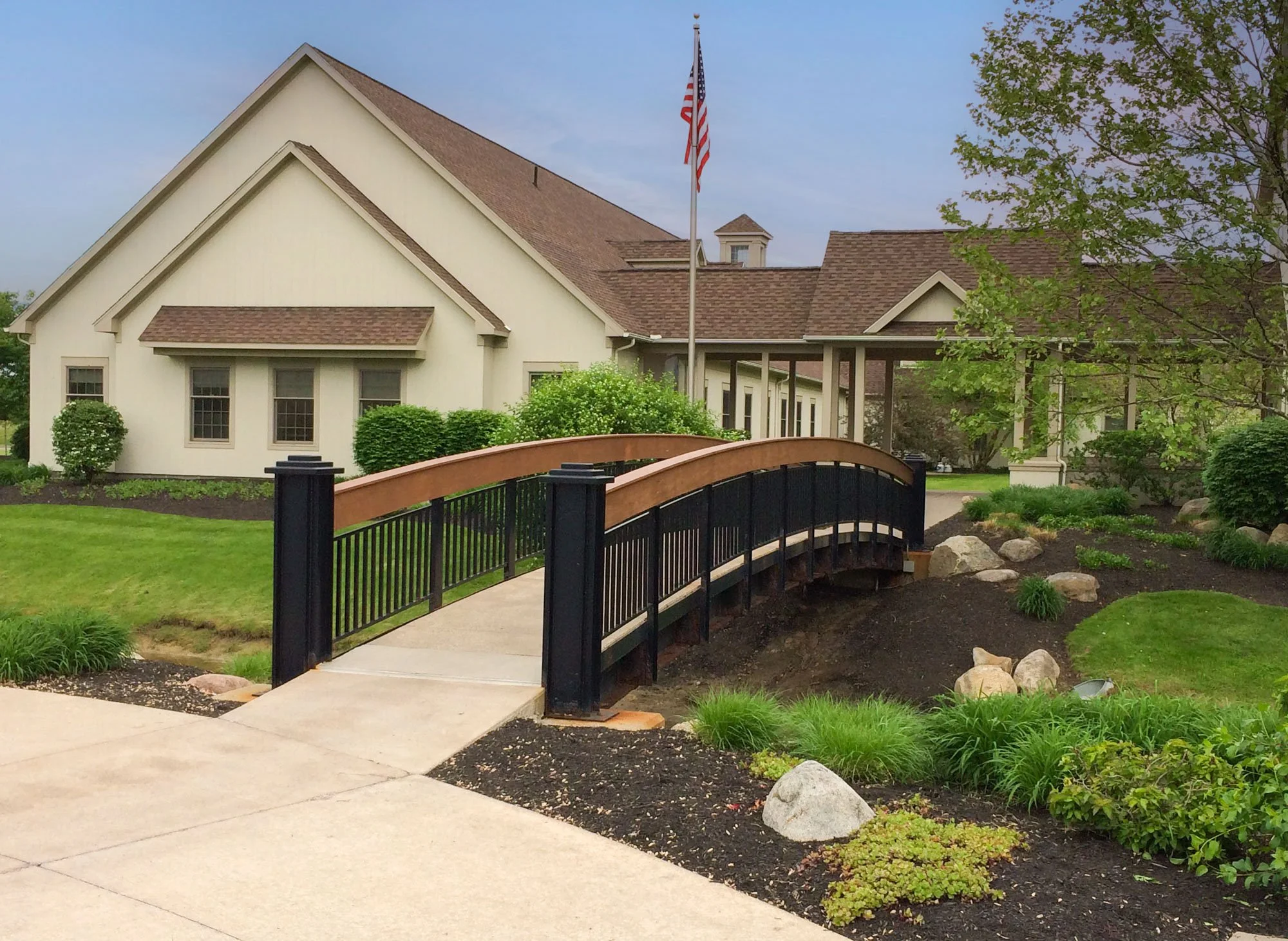 A white building with brown shingle roof, multiple windows, and a small tower with a flagpole displaying the American flag. There is a curved pathway, a black and wood railing, green trees, bushes, and landscaped garden beds in the foreground.