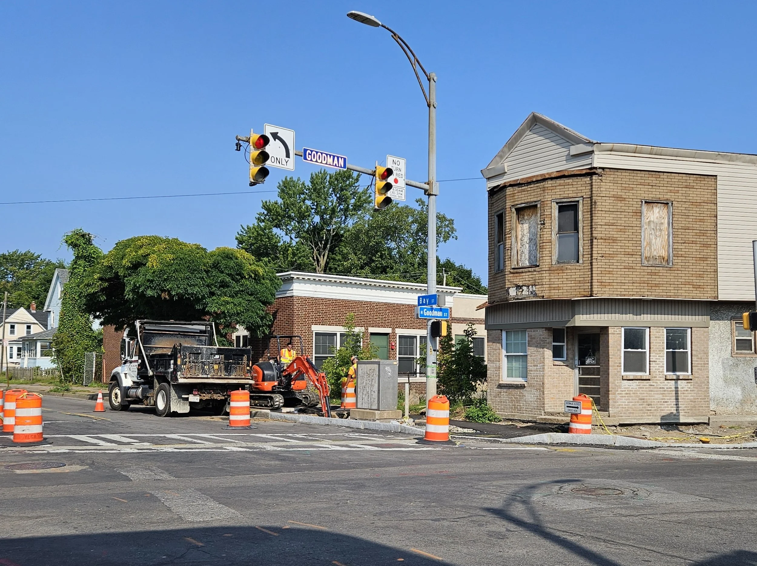 Street with construction work on the sidewalk, orange traffic cones, a construction vehicle, and a traffic light at an intersection under a clear blue sky.