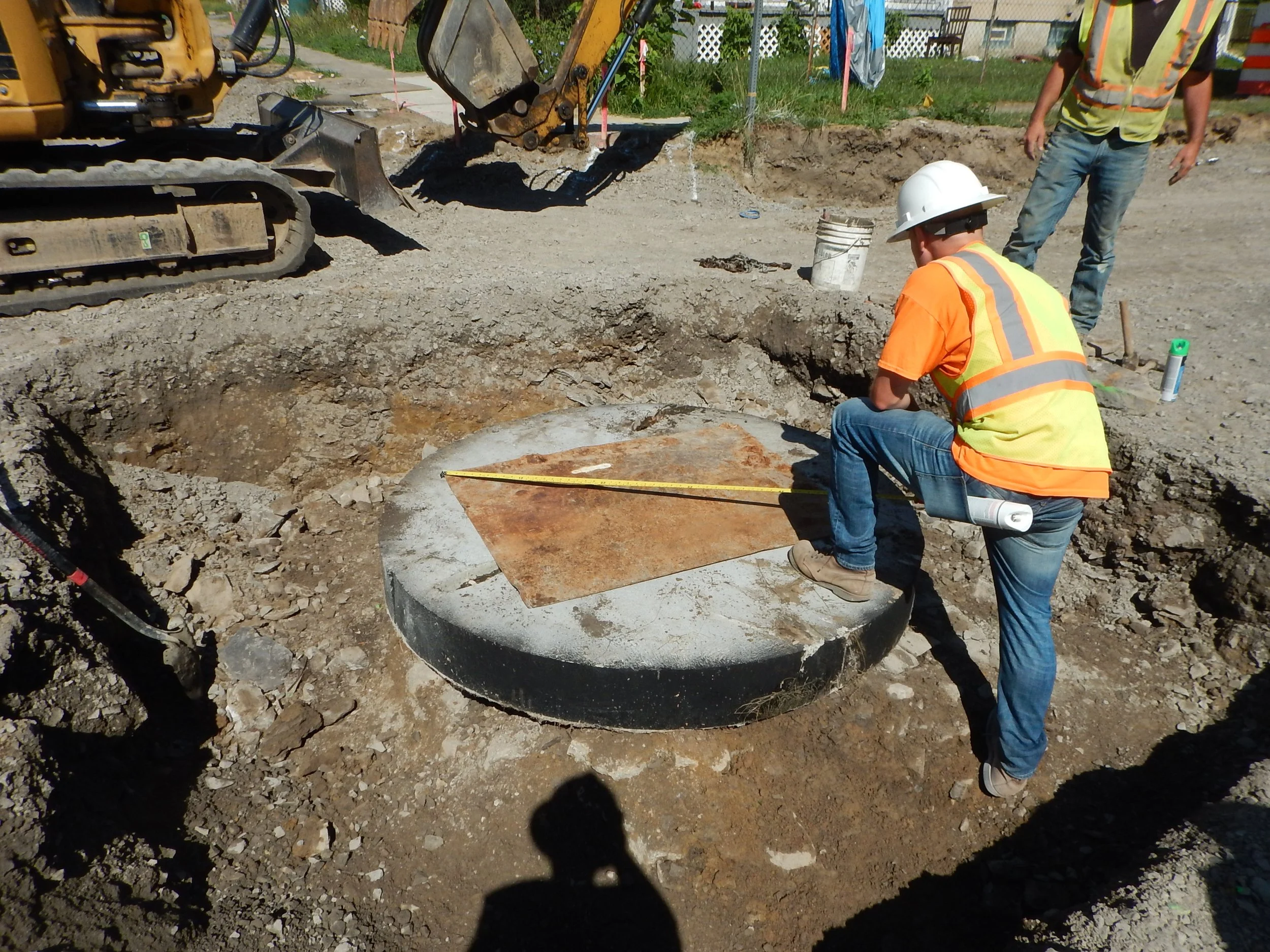 Construction workers at a site, one kneeling and another standing, near a large concrete structure and digging equipment. They are wearing safety vests and hats.