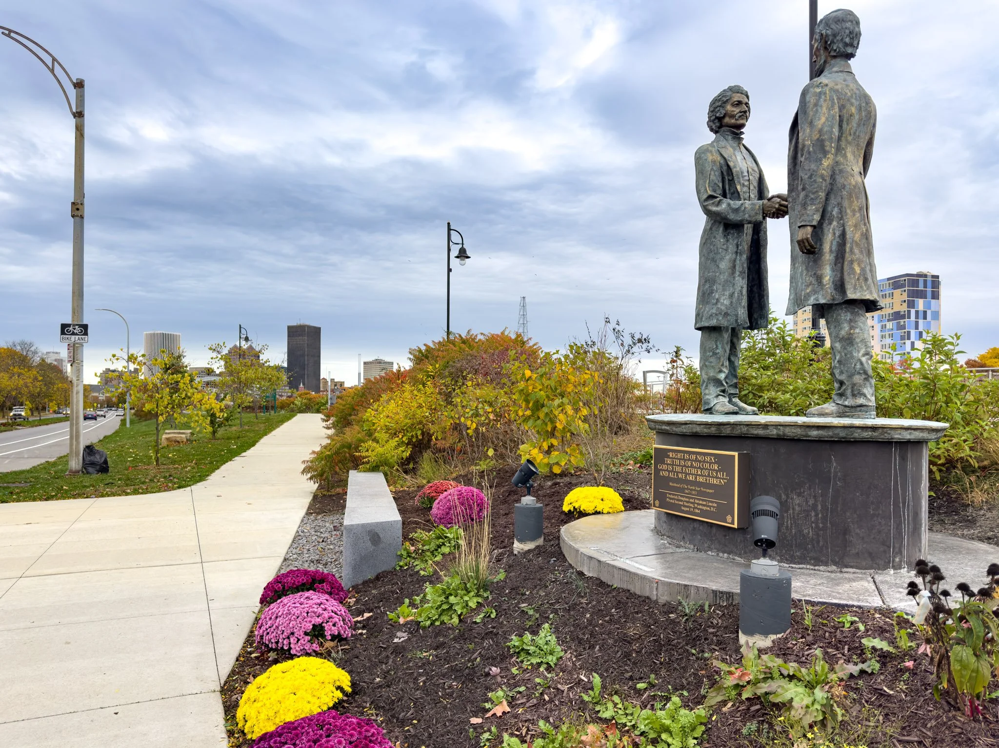 Statue of two men shaking hands, located in a city park with colorful flowers and trees, buildings and cloudy sky in the background.