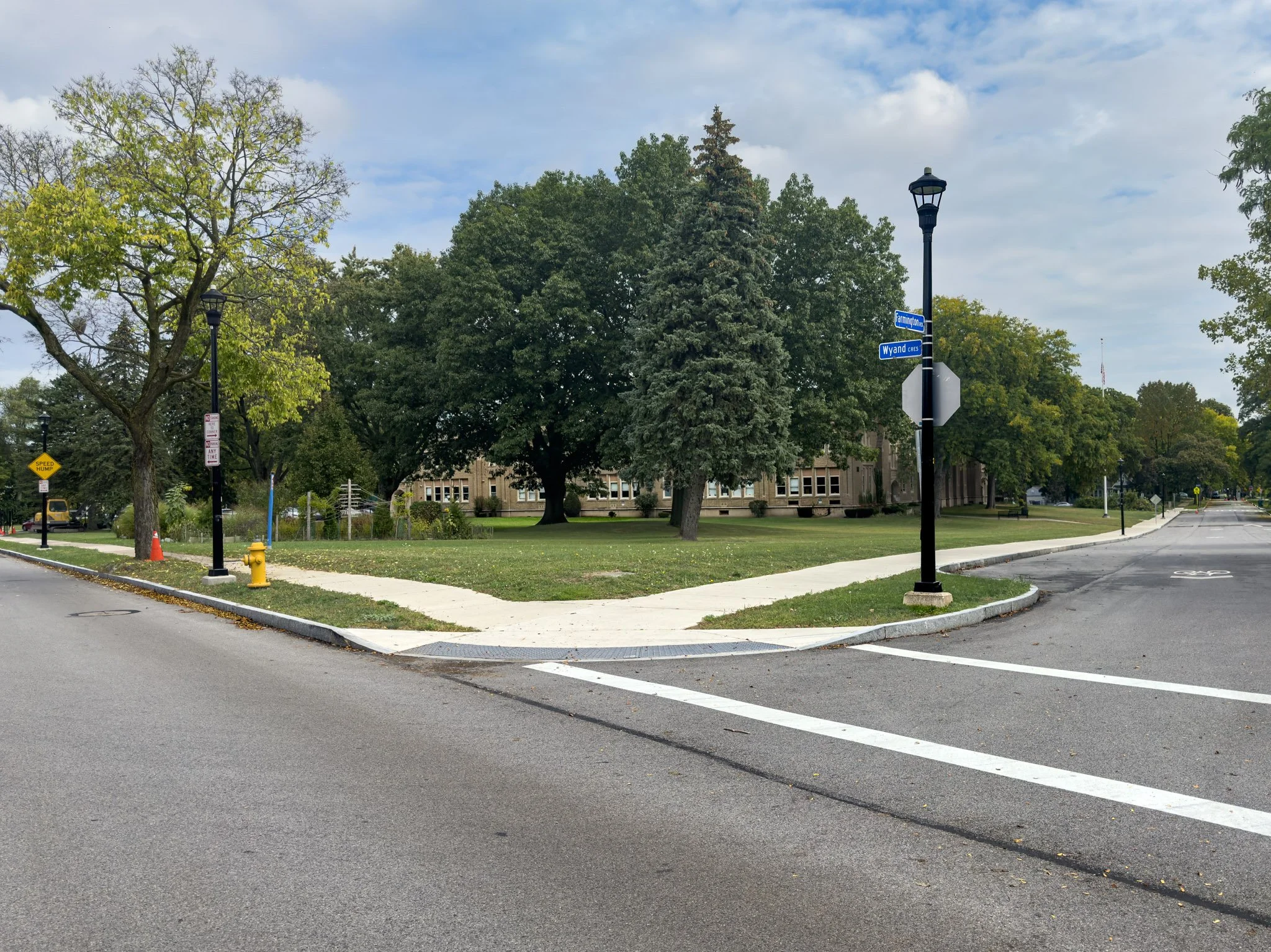 A residential street scene with a sidewalk, grass, trees, street signs, a fire hydrant, and a building in the background. The sky is partly cloudy.
