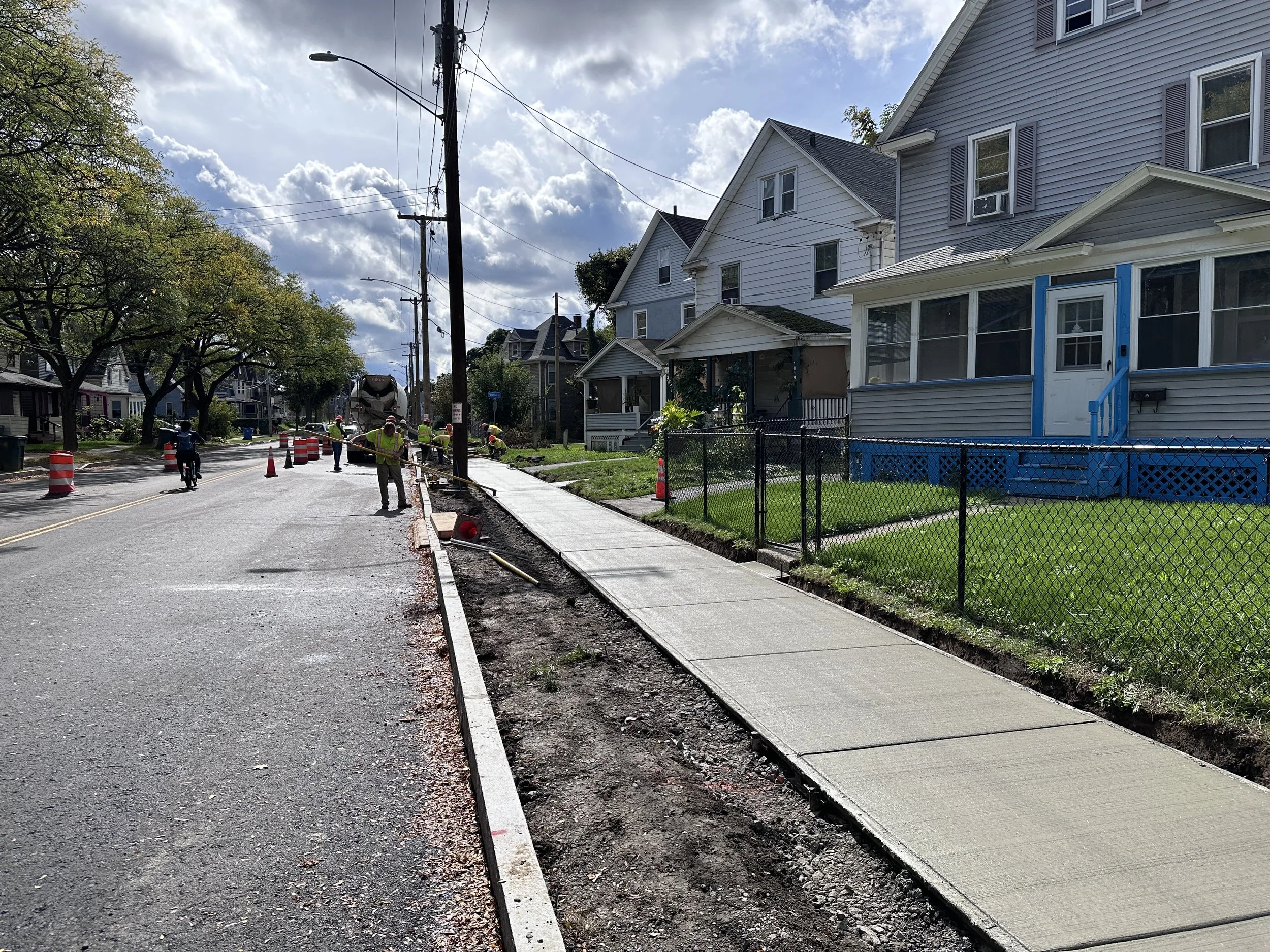 Street with sidewalk under construction, construction workers working on the sidewalk, houses on the right side, trees on the left side, sky with clouds.