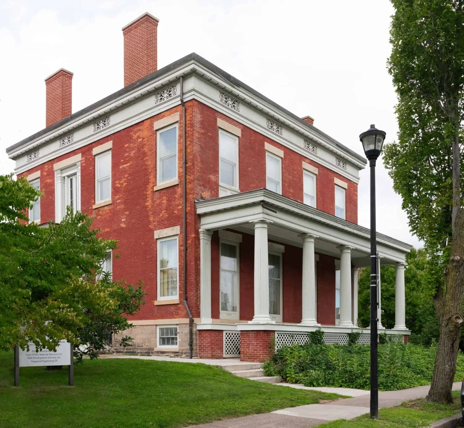 The Vanguard Engineering office, a three-story red brick building with white columns and a porch, surrounded by green trees and grass, with a sidewalk and streetlamp in front.