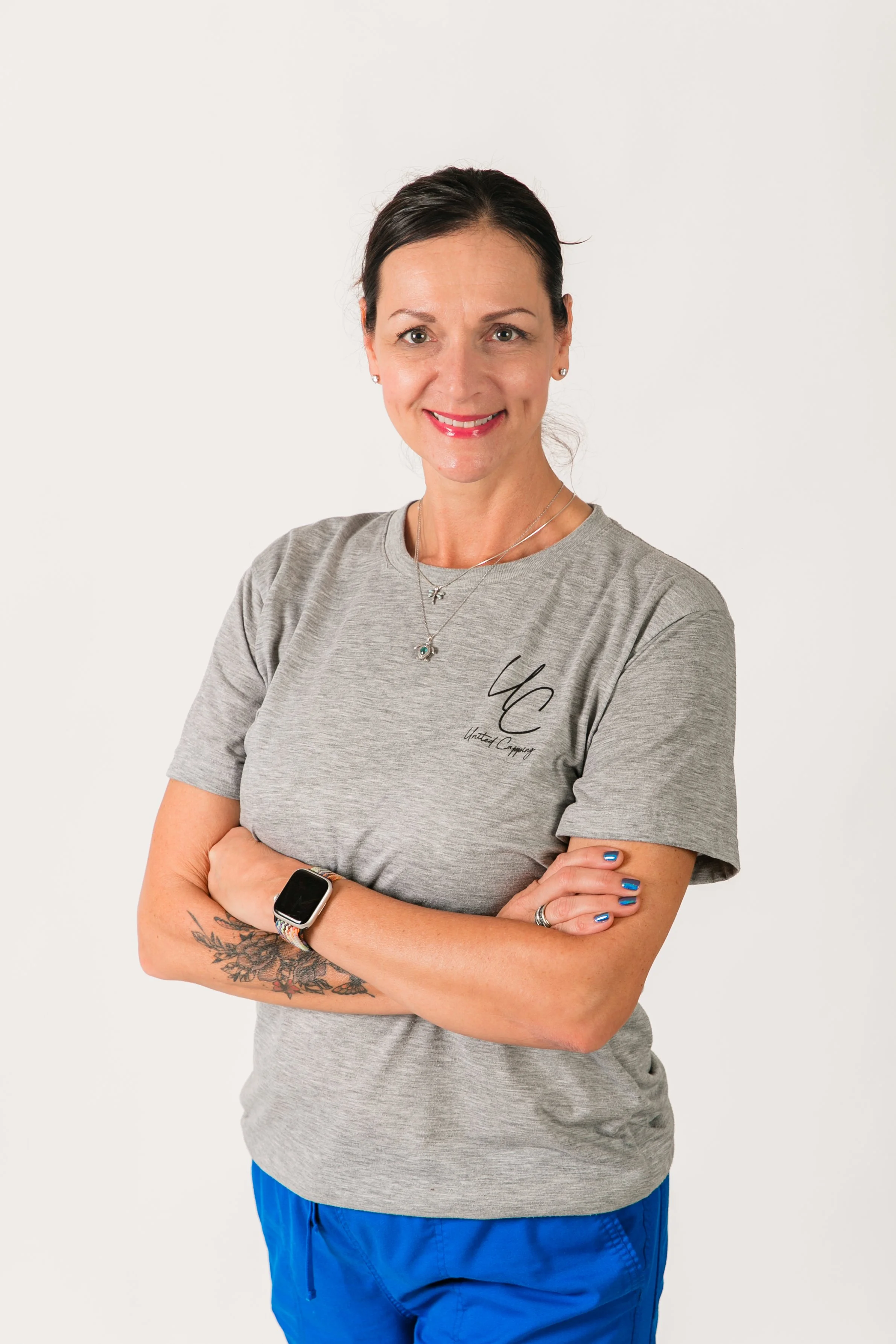 A woman with dark hair pulled back, smiling, wearing a light gray t-shirt with black text and blue athletic shorts, standing against a plain white background.