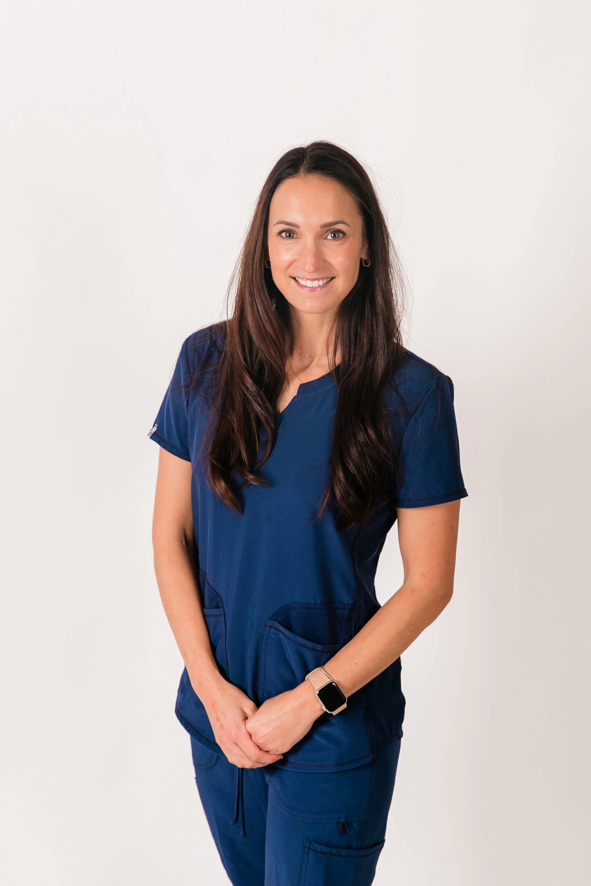 A woman in dark blue medical scrubs standing against a plain white background smiling with her hands clasped in front of her. She has long dark hair, earrings, and is wearing a smartwatch.