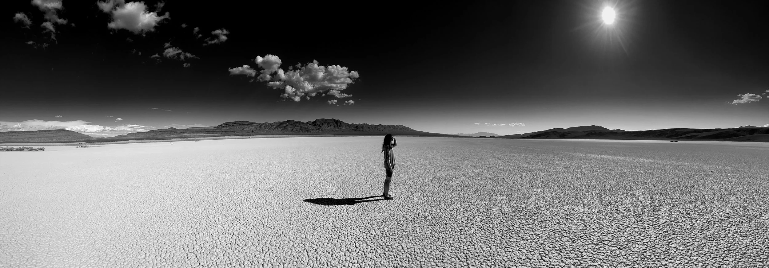 A black and white photo of a person standing alone in a vast, dry desert landscape with cracked ground, mountains on the horizon, clouds in the sky, and the sun shining.