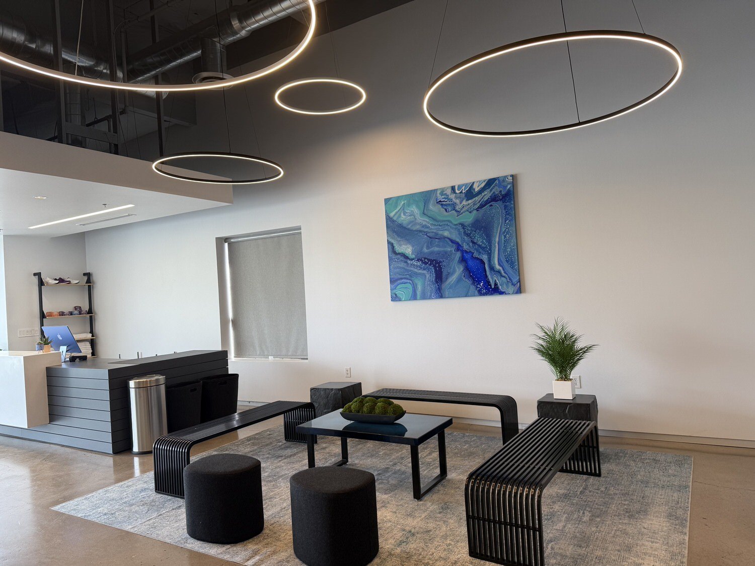 Modern office lobby with circular ceiling lights, abstract blue artwork, gray check-in counter, black benches and stools, and a small potted plant.