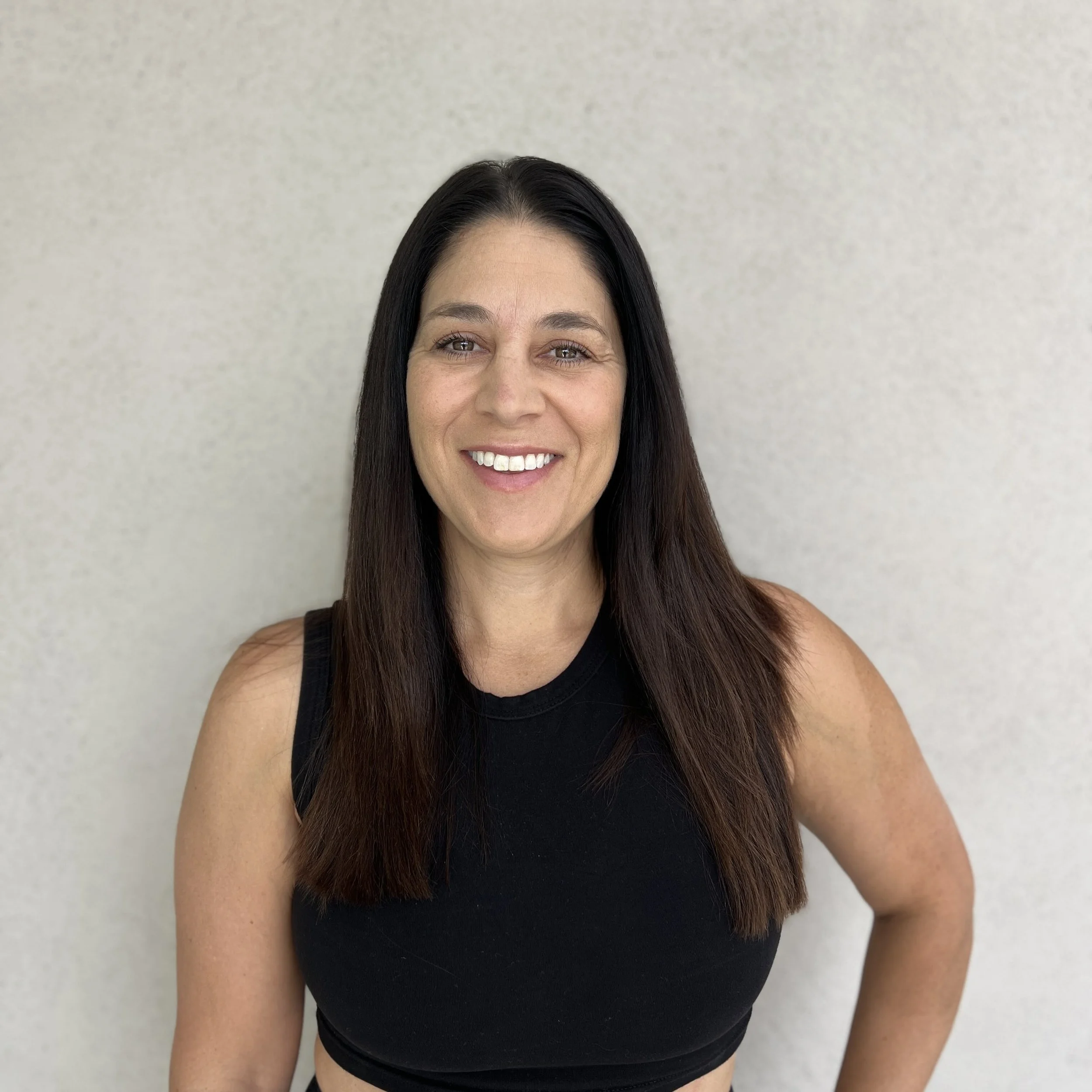A woman with long dark hair smiling while wearing a black sleeveless top standing against a plain light-colored wall.