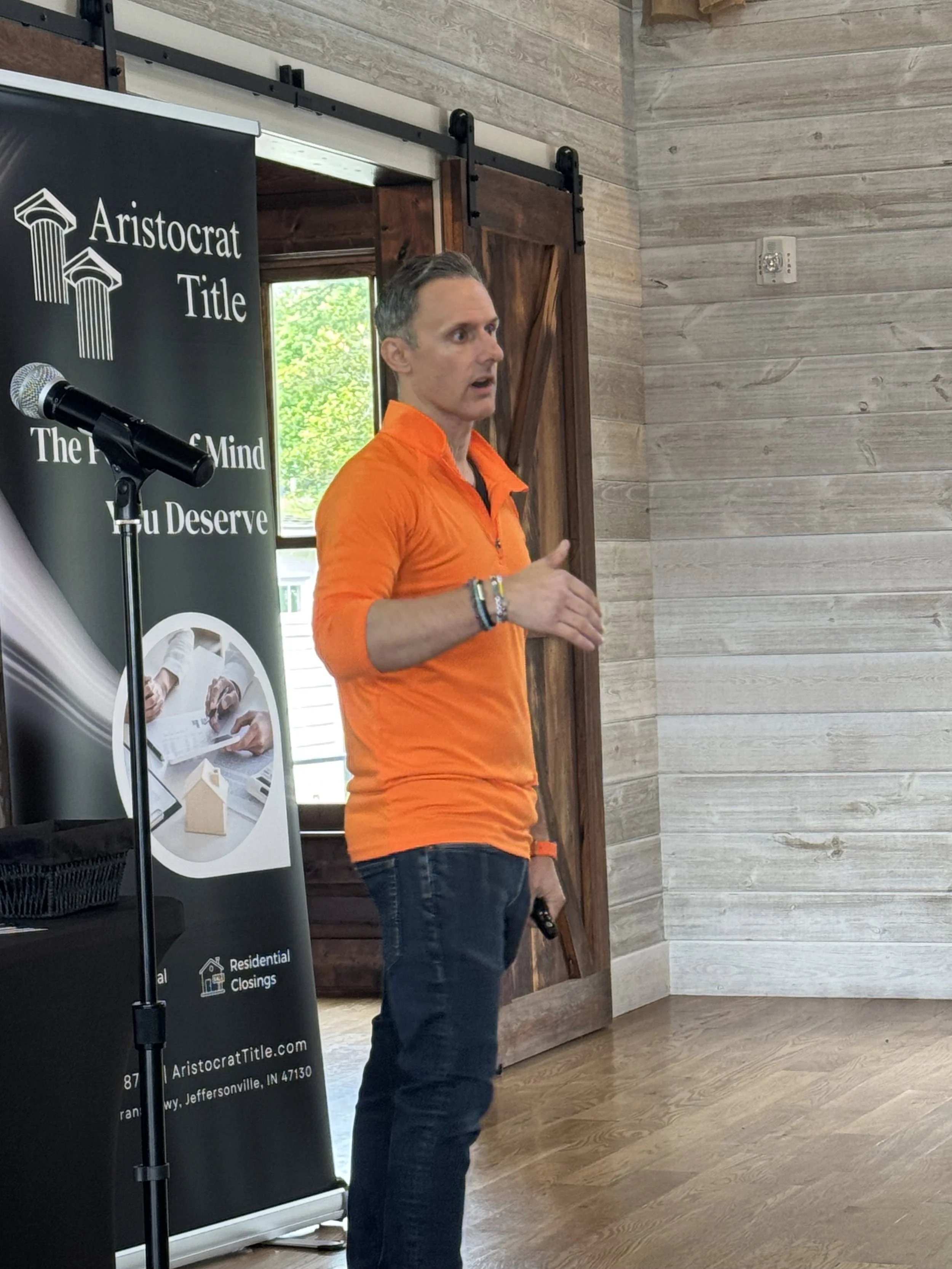 A man in an orange shirt giving a presentation indoors, standing near a black banner with the words "Aristocrat Title" and a microphone on a stand.