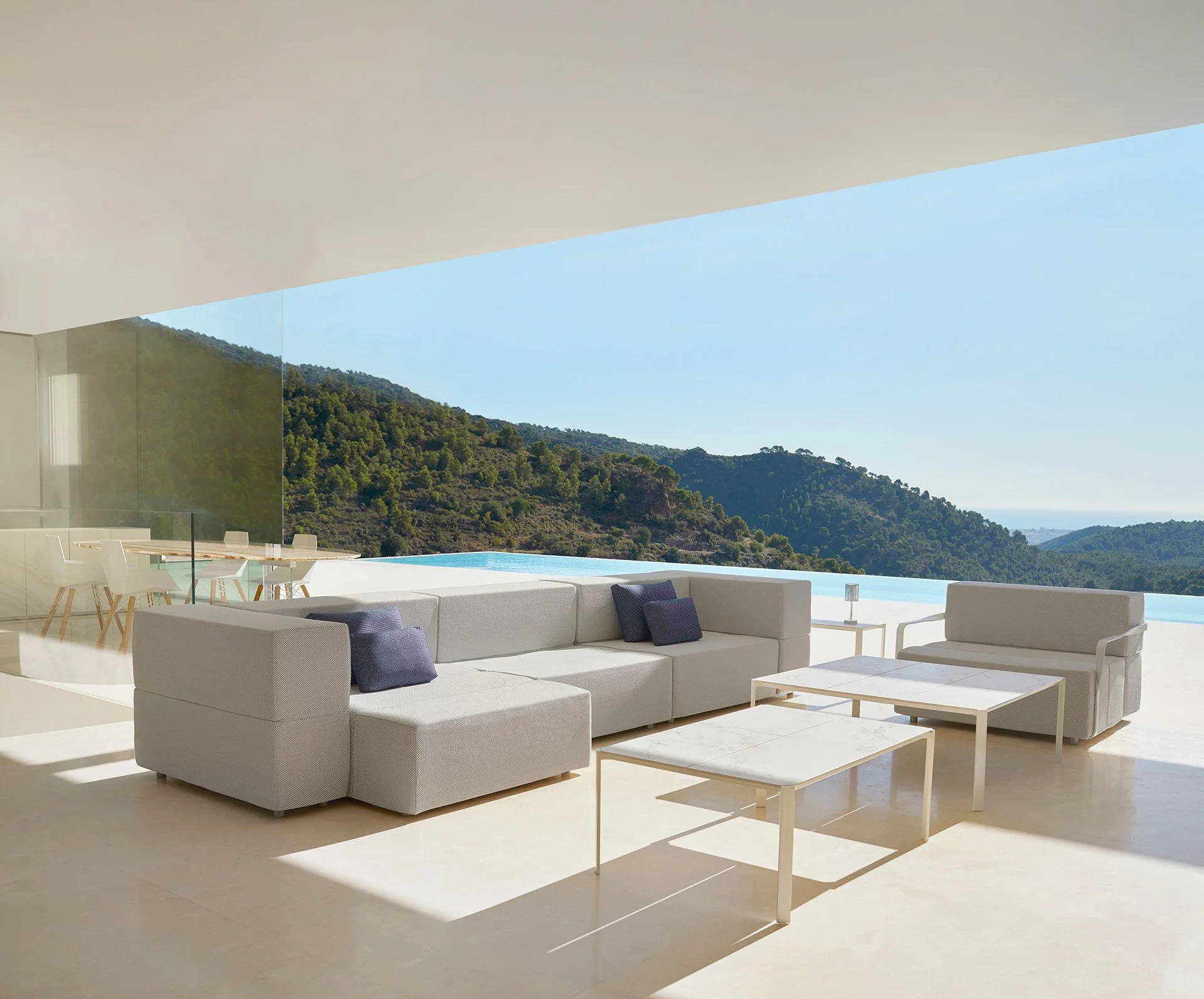 Sala moderna con sillones blancos y mesas de café en un espacio con vista a montañas verdes y cielo despejado