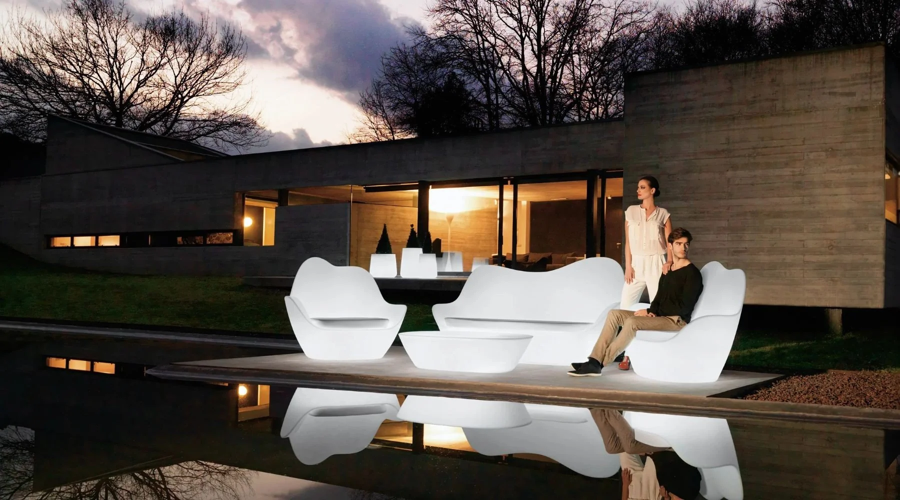 Muebles de diseño blanco en un patio de noche con una casa moderna de fondo, reflejados en un cuerpo de agua