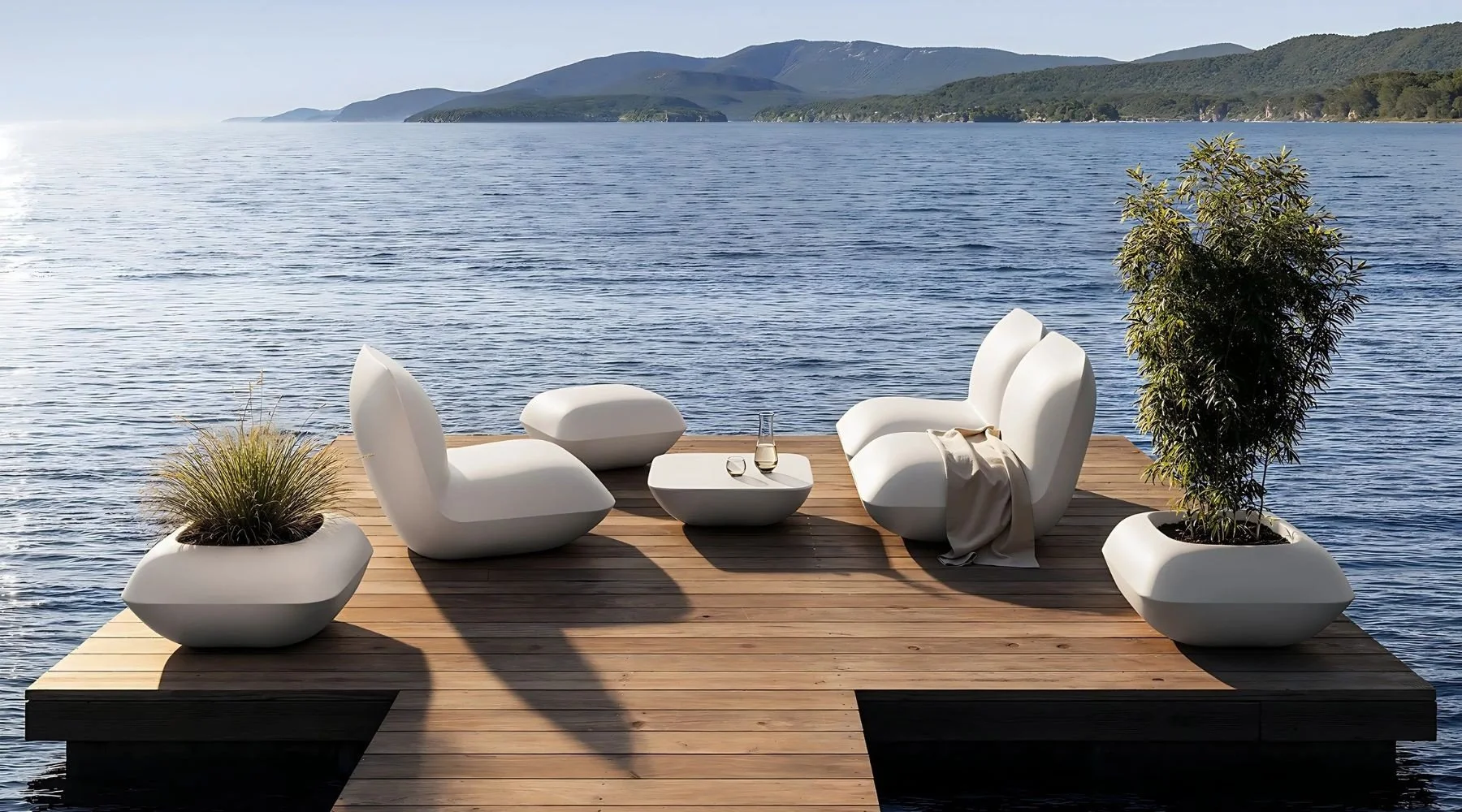Muebles de diseño moderno en una terraza sobre el agua, con vista a montañas y un lago, incluyendo sillones blancos, una mesa central y plantas en macetas blancas.