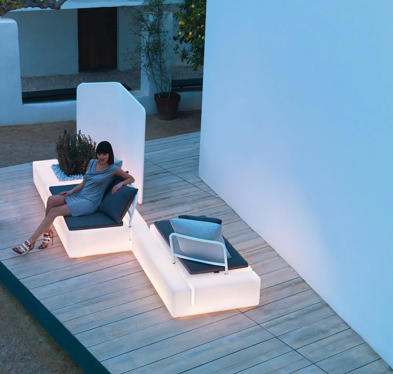 Mujer sentada en mobiliario de exterior con iluminación suave en un entorno moderno y minimalista al atardecer.