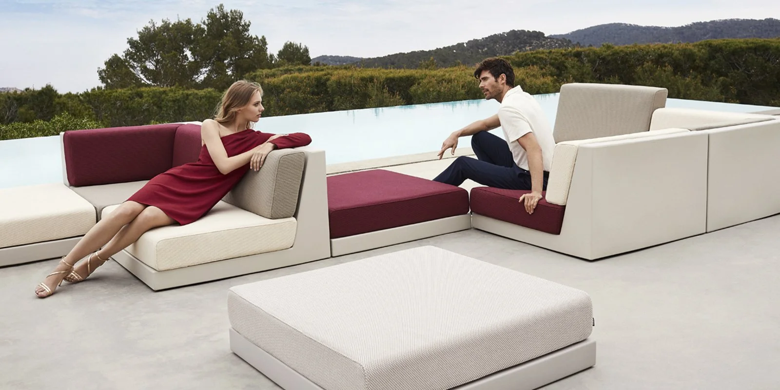 Una pareja sentada en un sillón modular en una terraza con vista a un paisaje natural y una piscina infinita en el fondo.