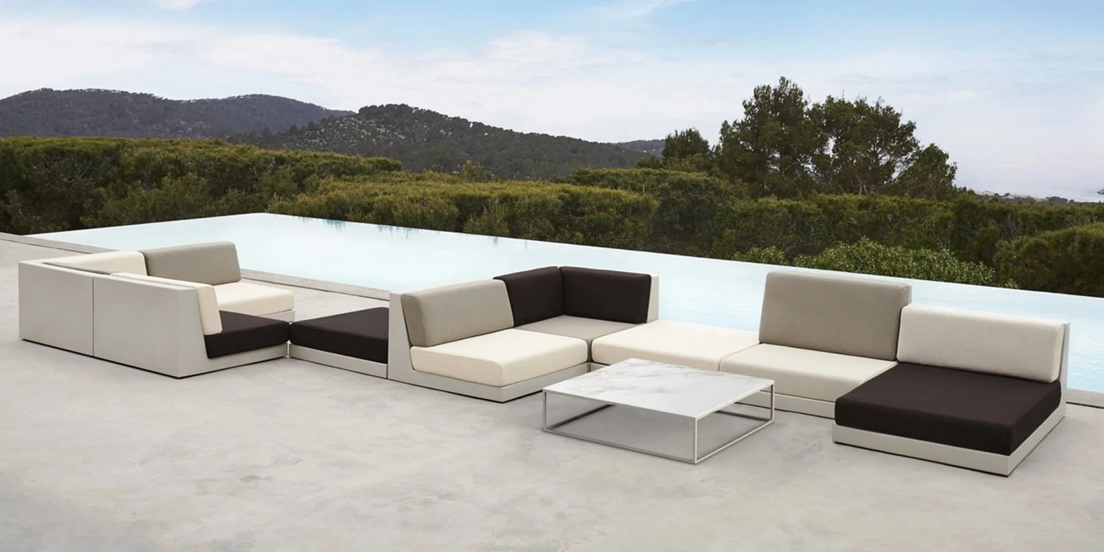 Conjunto de sillones modernos blancos y oscuros junto a una mesa de mármol en una terraza con vista al campo y montañas en el fondo.