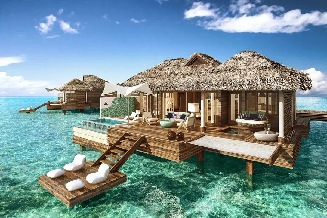 Villa sobre el agua con techo de paja, área de descanso con sillones, sombrillas y piscina pequeña, rodeada de agua cristalina y cielo despejado.
