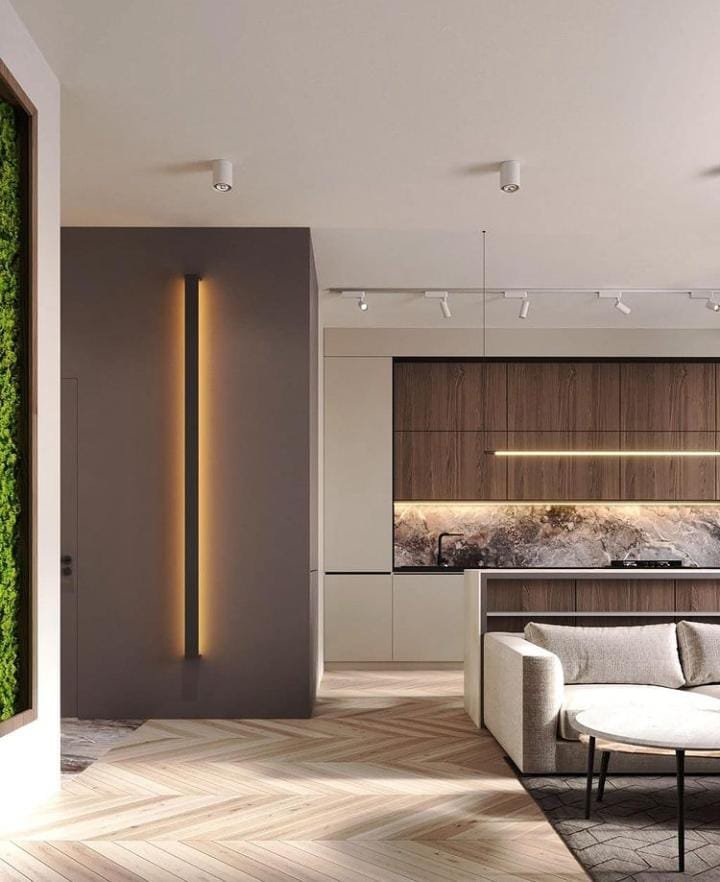Sala moderna con diseño minimalista, iluminación empotrada en paredes y techo, decoración con panel de vegetación vertical, pared de acento con tono gris y cocina integrada con muebles de madera y encimera de mármol. Sofá claro y mesa de centro redonda sobre alfombra gris.