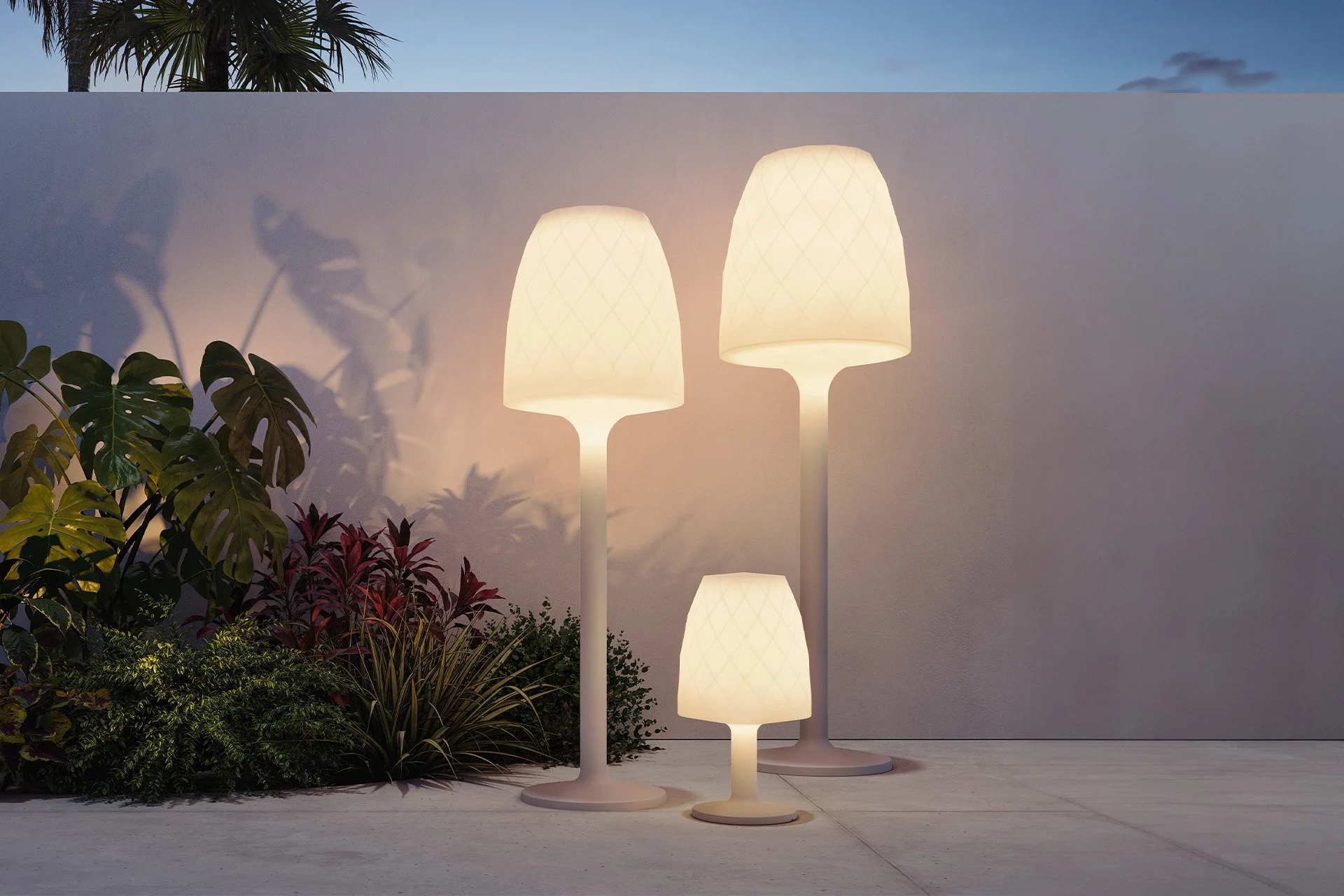 Tres lámparas de pie de color blanco con pantallas que emiten luz cálida, ubicadas junto a plantas verdes y rojas en un espacio exterior durante el atardecer.