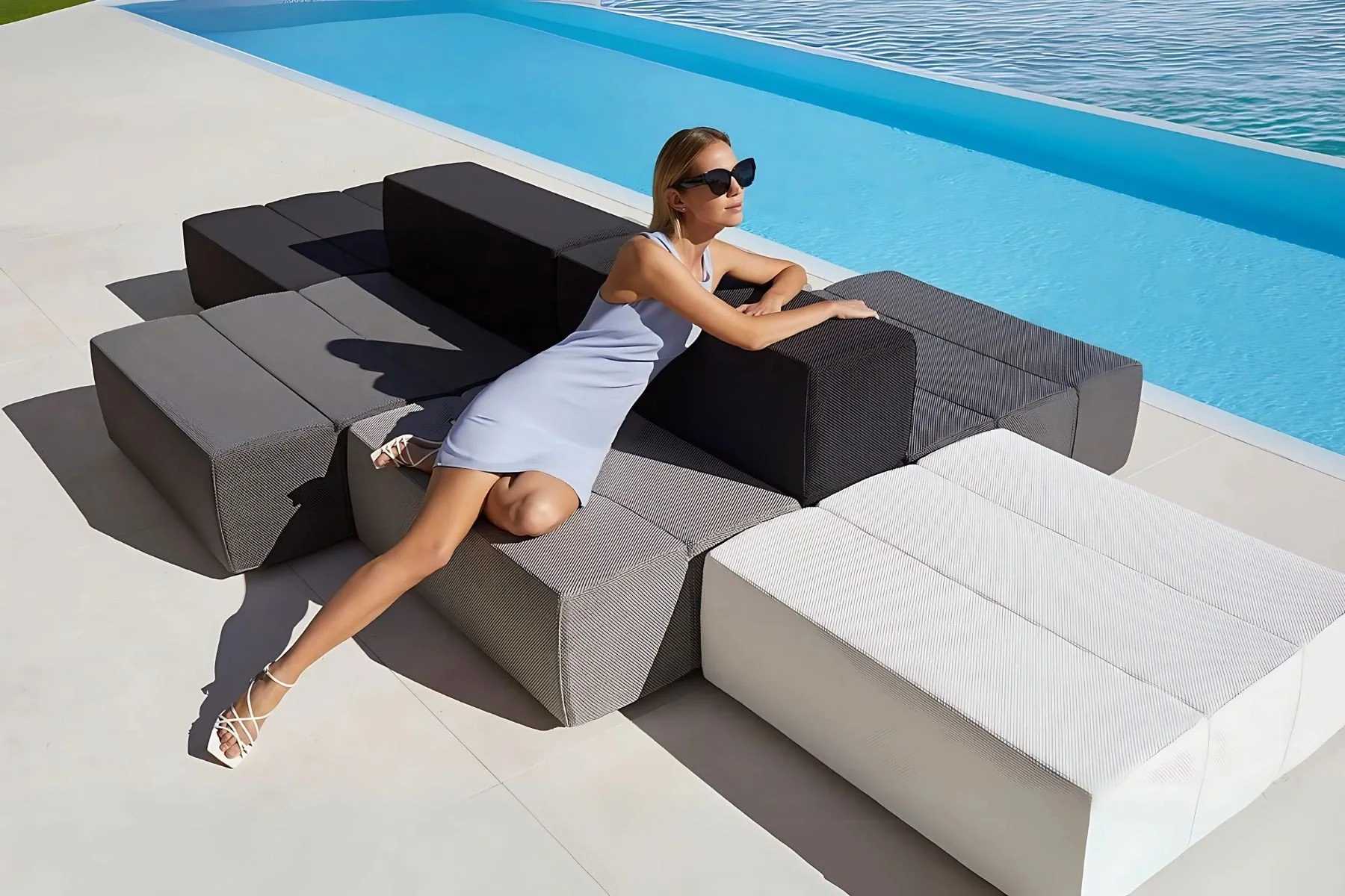 Mujer descansando en muebles de exterior junto a una alberca, con gafas de sol y vestido blanco.