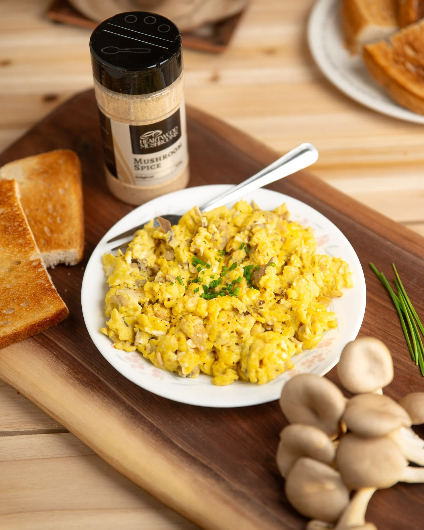 Scrambled eggs with green chives, a jar of Heartwood Mushrooms mushroom spice in flavor 'original,' toasted bread, a bunch of garlic, and a plate of baked goods on a wooden table.