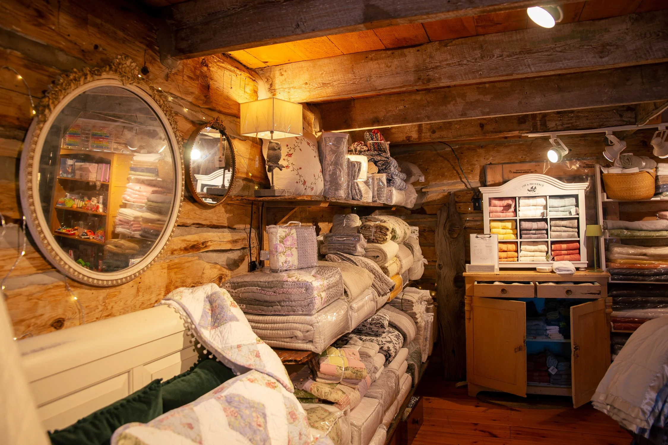 Interior of a cozy bedding shop with stacked folded linens, pillows, and blankets on rustic wooden shelves. Two circular mirrors are hung on the wooden wall, with soft warm lighting creating a welcoming atmosphere.