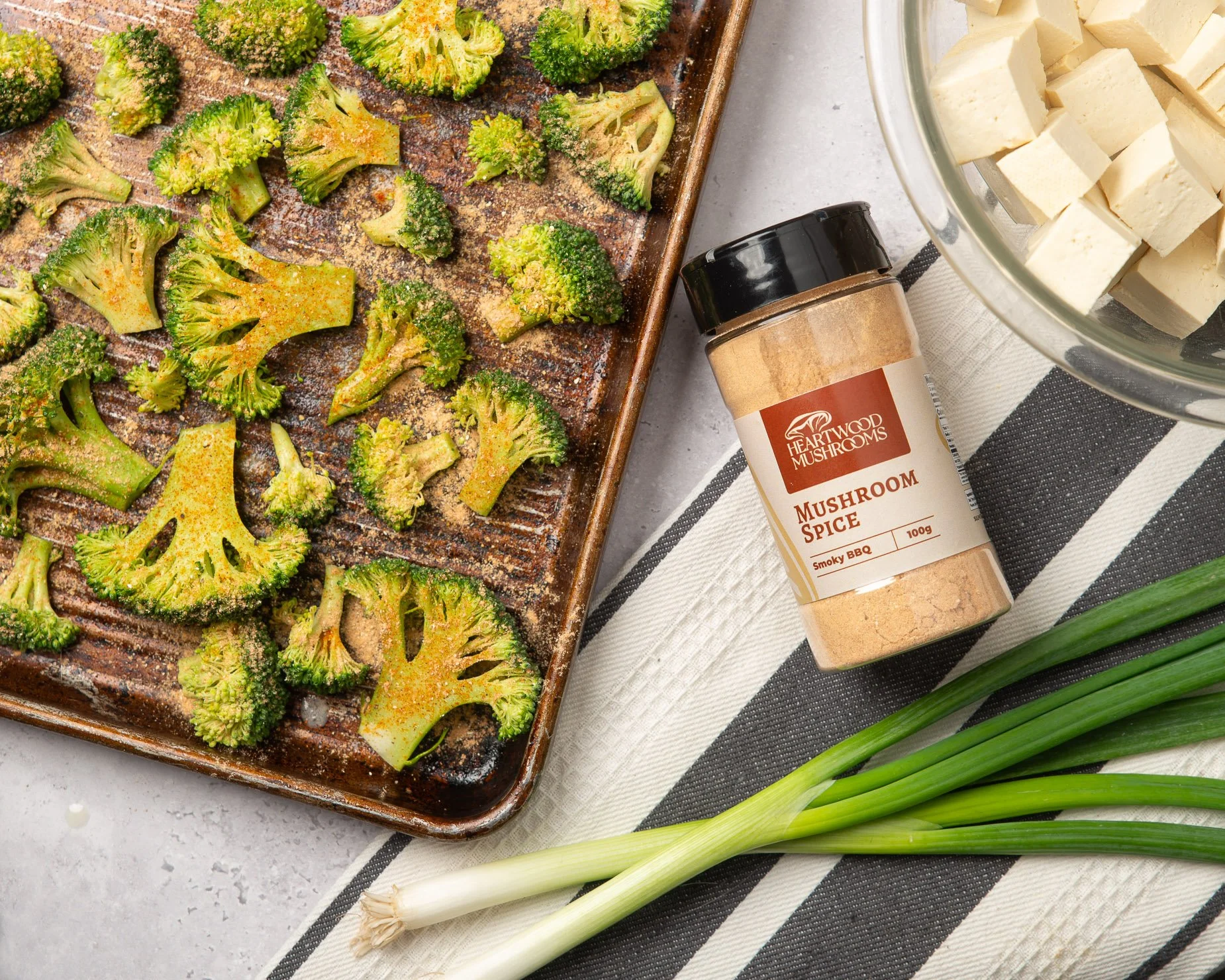 Roasted broccoli on a baking sheet with seasoning, a container of vegan tofu with cubed tofu, a jar of mushroom spice labeled 'Smoky BBQ' from Heartwood Mushrooms, green onions, and a bowl of seasoning.