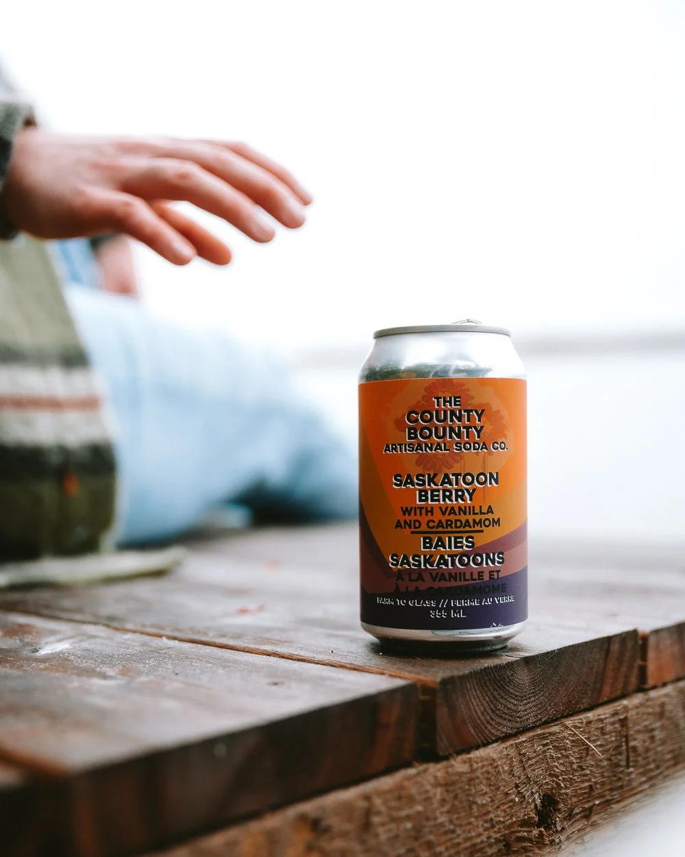 A can of Saskatoon Berry soda with vanilla and cardamom flavor from The County Bounty Artisan Soda Co. placed on a wooden table, with a hand reaching toward it.