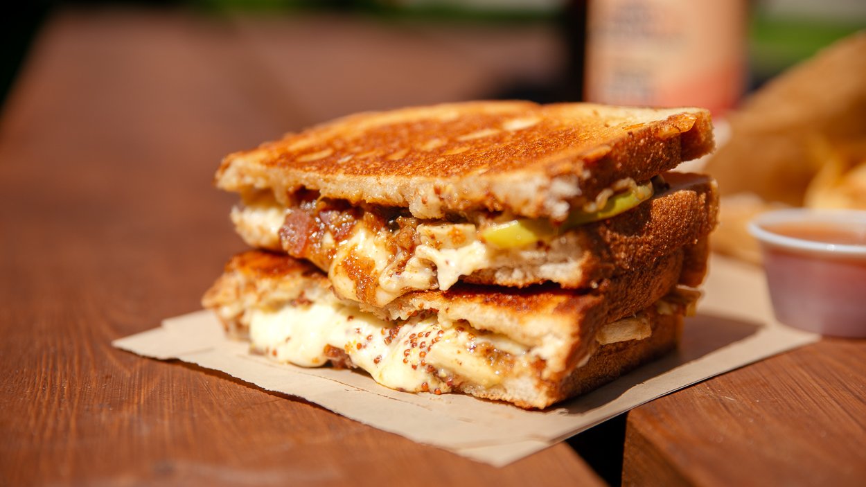 Close-up of a toasted grilled cheese sandwich with bacon, dijon mustard, and apple slices on a wooden table, with a small cup of dipping sauce, homemade chips in a paper bag, and a canned drink in the background.
