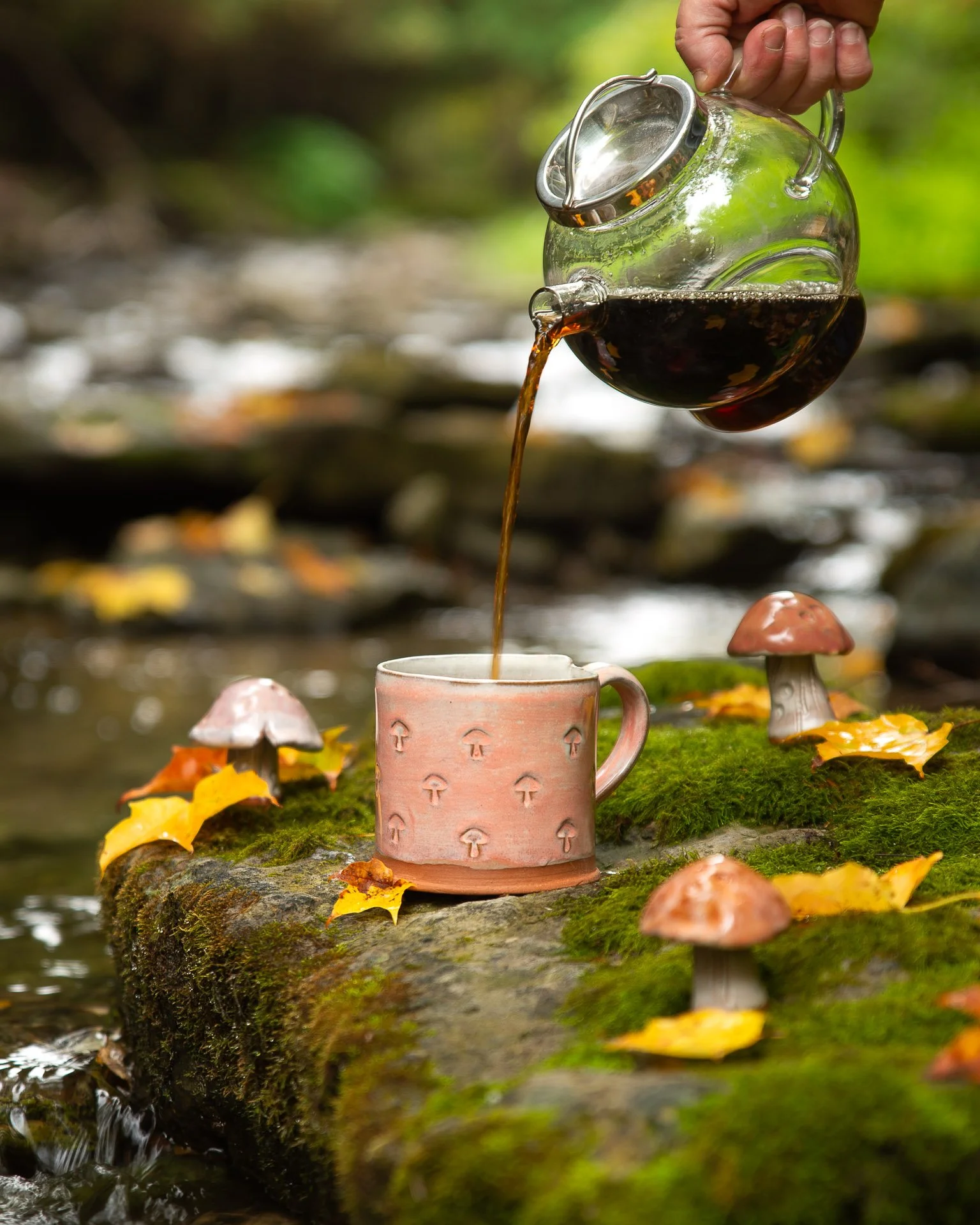 Coffee or tea being poured into a pink mug with mushroom decorations on it, placed on a moss-covered log or rock surrounded by autumn leaves and small ceramic mushrooms in a forest stream setting.
